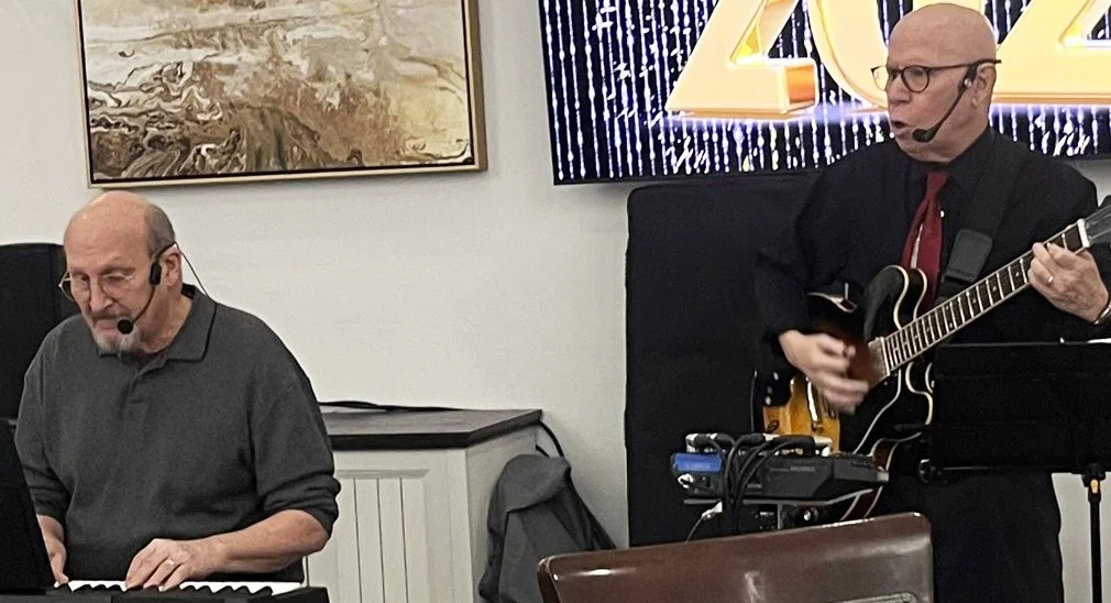 Two musicians performing indoors; one playing keyboard and the other playing guitar, both wearing headsets.