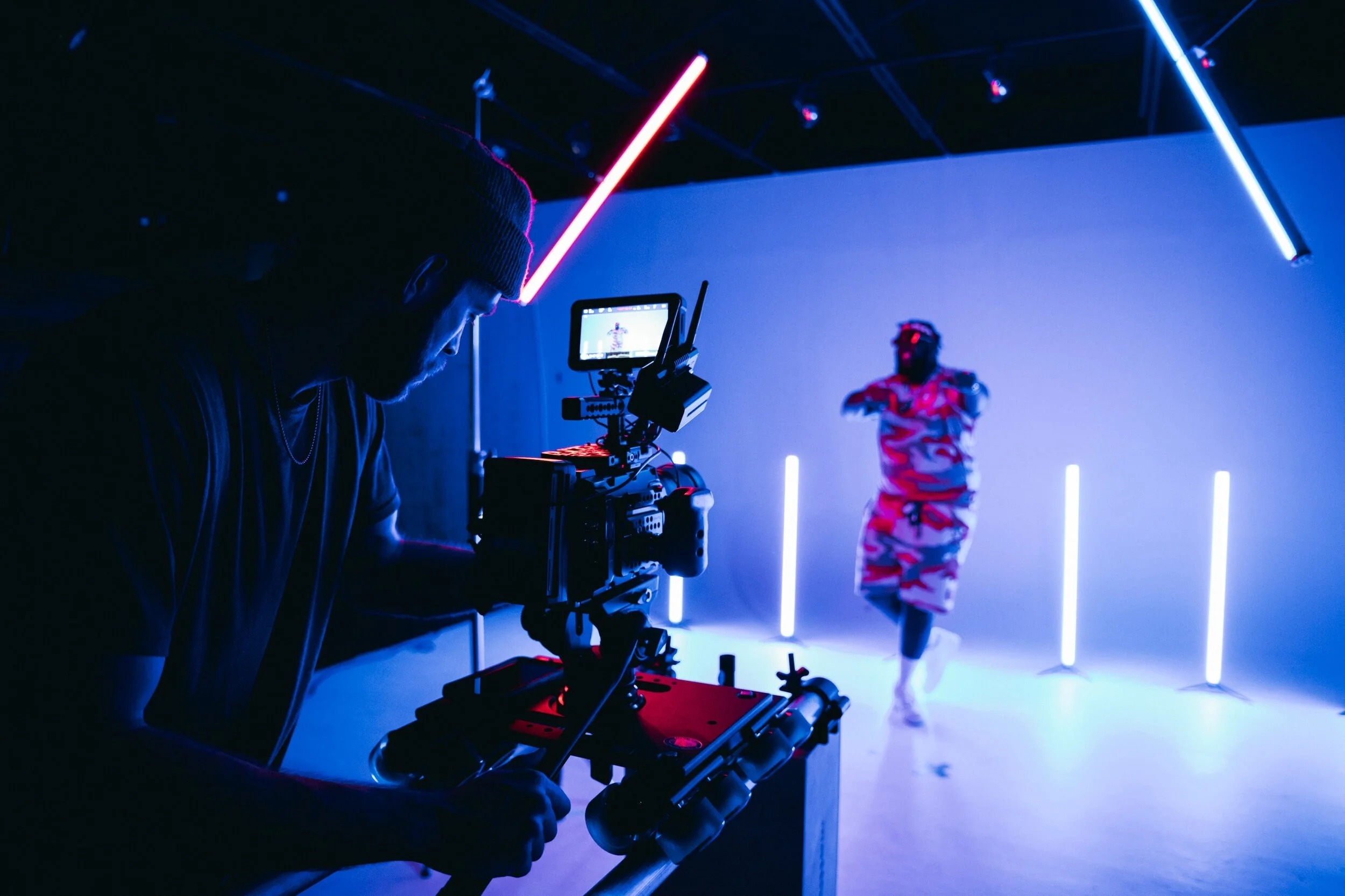 A person is filming a dancer in a dark, modern studio with blue and pink neon lighting.