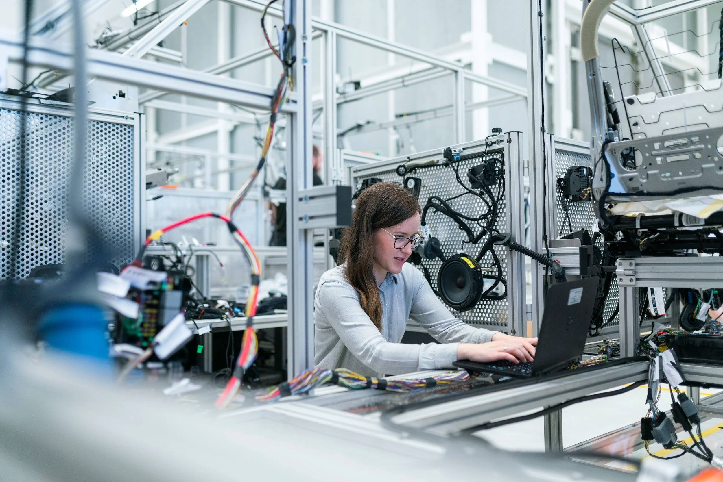 A woman wearing glasses working on a laptop in an industrial setting filled with electronic equipment, wires, and metal frames.
