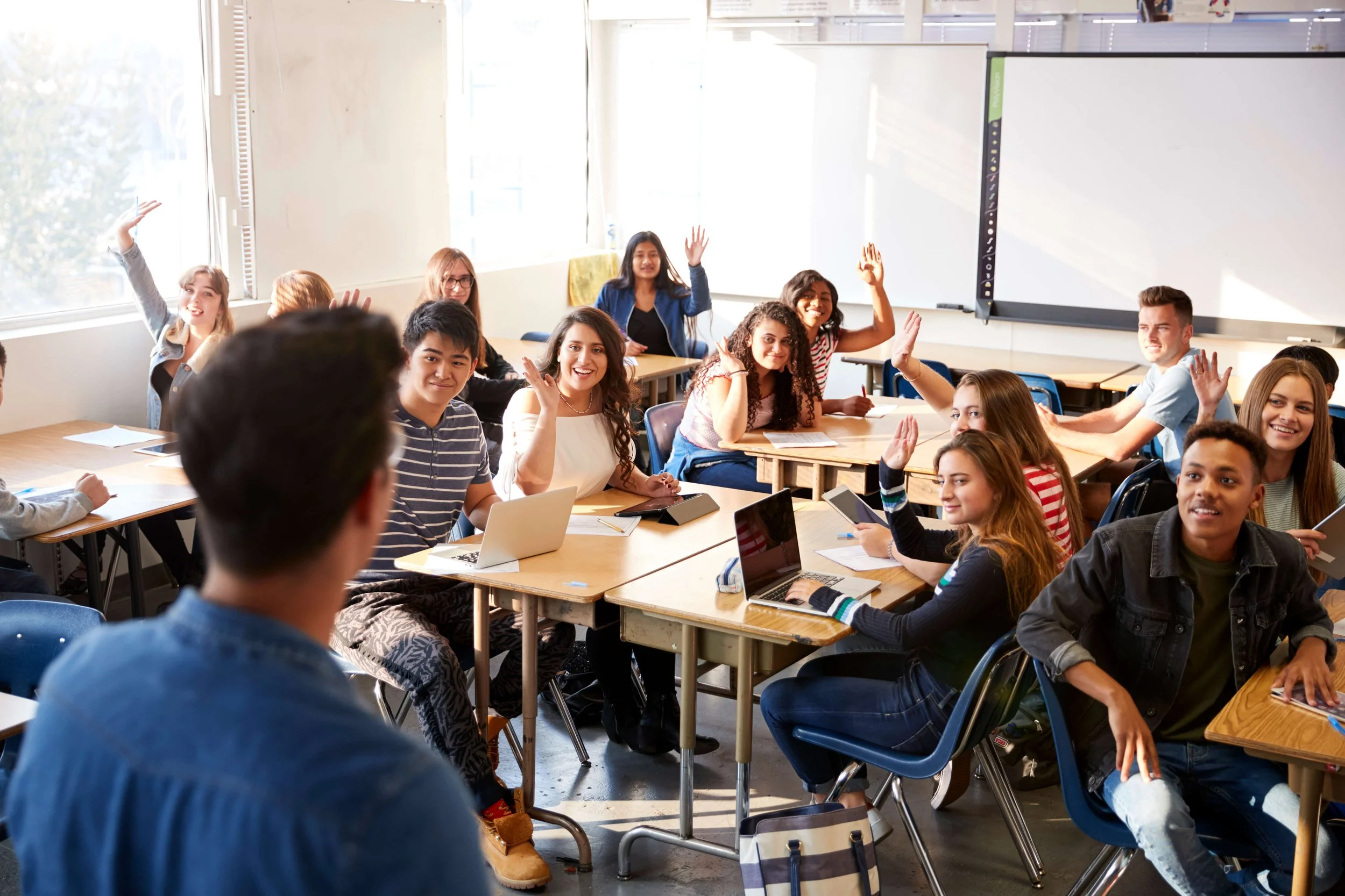 A classroom filled with students sitting at desks, some raising hands, and a teacher at the front facing the students.
