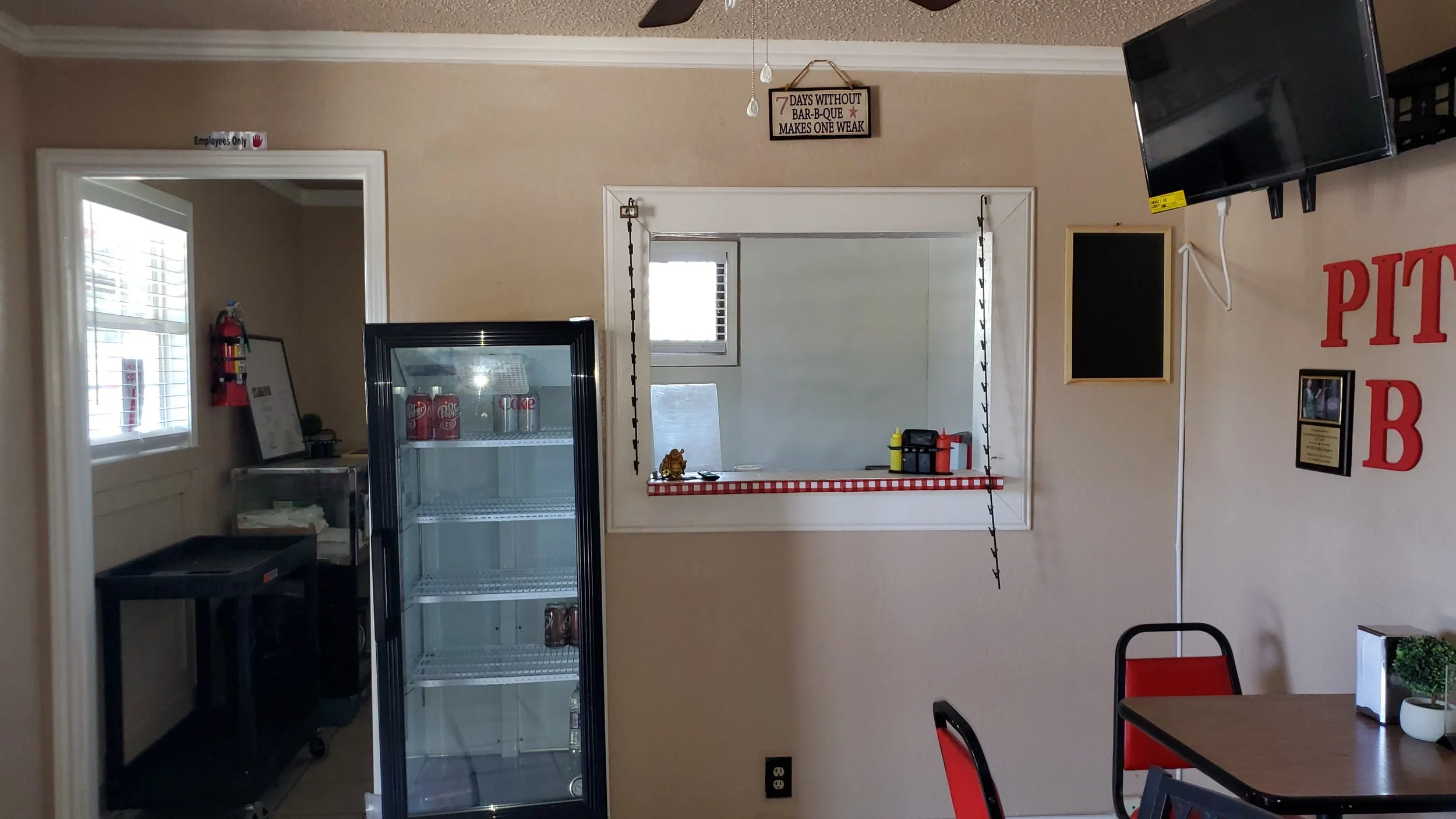 Interior of a small restaurant with a drink fridge, ordering window, red chairs, and a black TV on the wall.