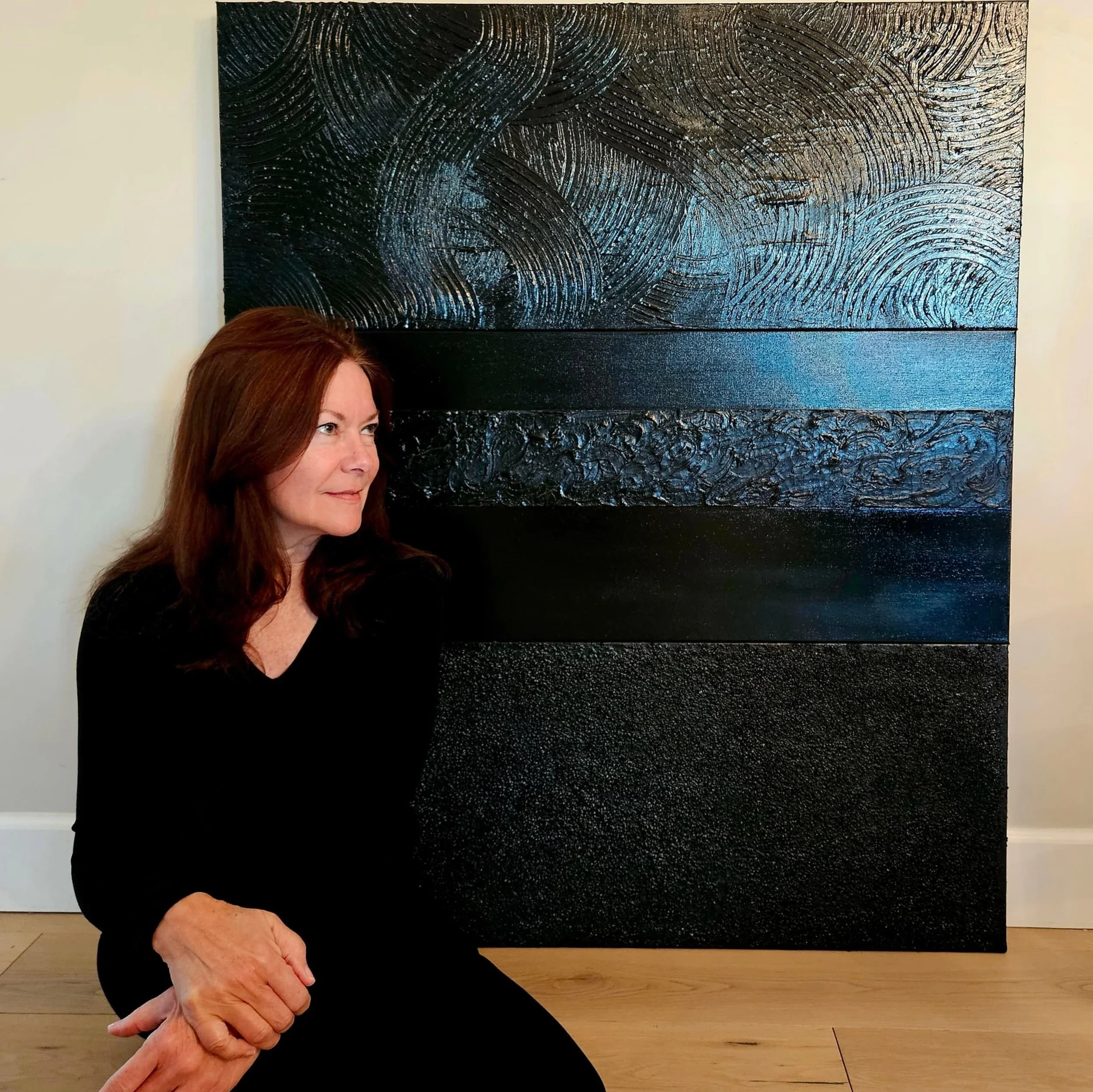 A woman with reddish-brown hair sitting on the floor in front of a large abstract black and blue textured painting.