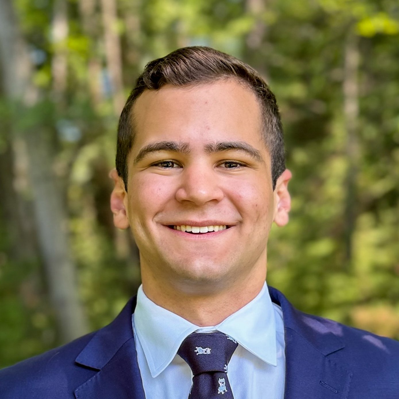 A young man in a suit and tie smiling outdoors with blurred trees in the background.