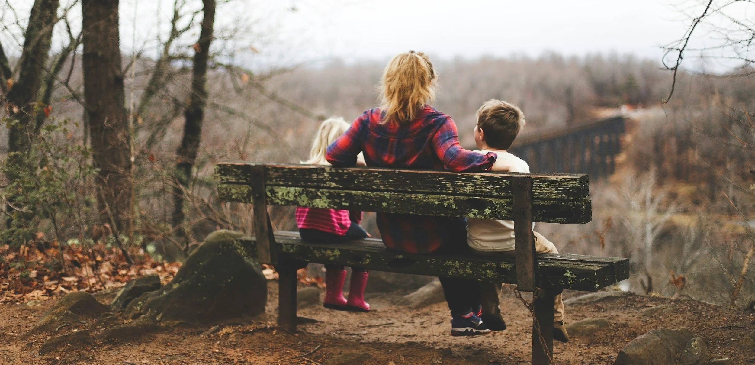 A woman with red hair in a plaid shirt sitting on a weathered wooden bench with two children in a wooded area during autumn, with a train passing on a distant bridge in the background.