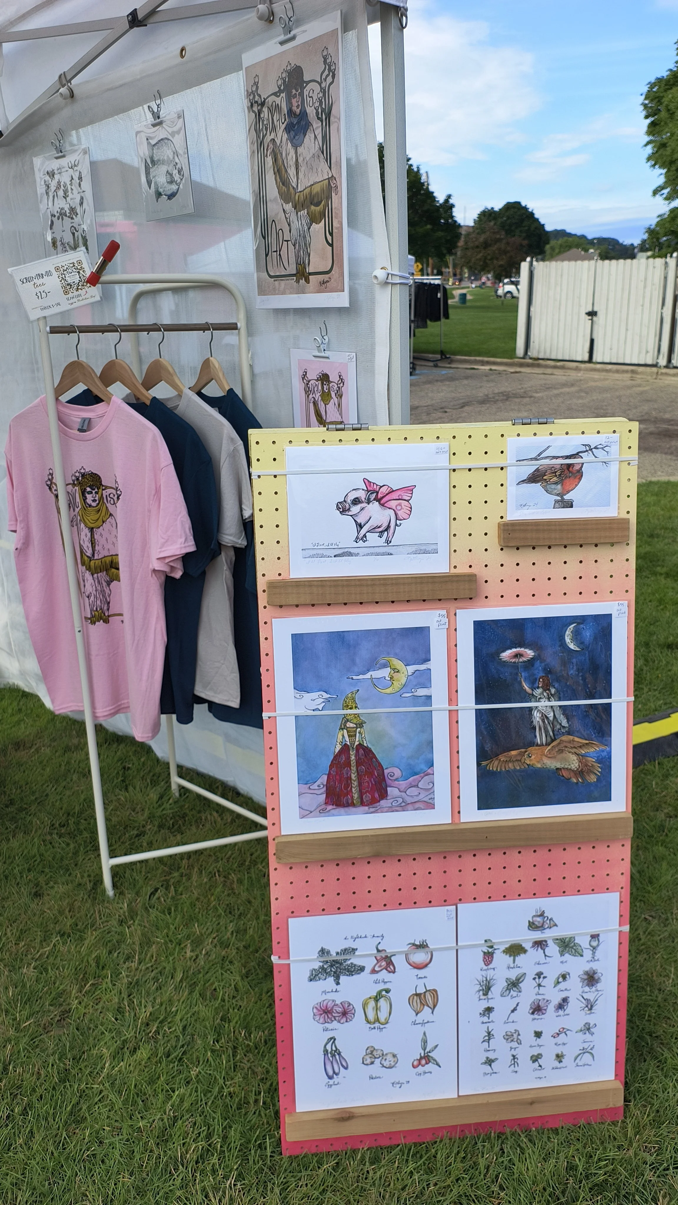 Photograph of the Artsy Rooster Studio's Fair Booth, with Drag is Art tees, and illustration prints on a colorful rack.