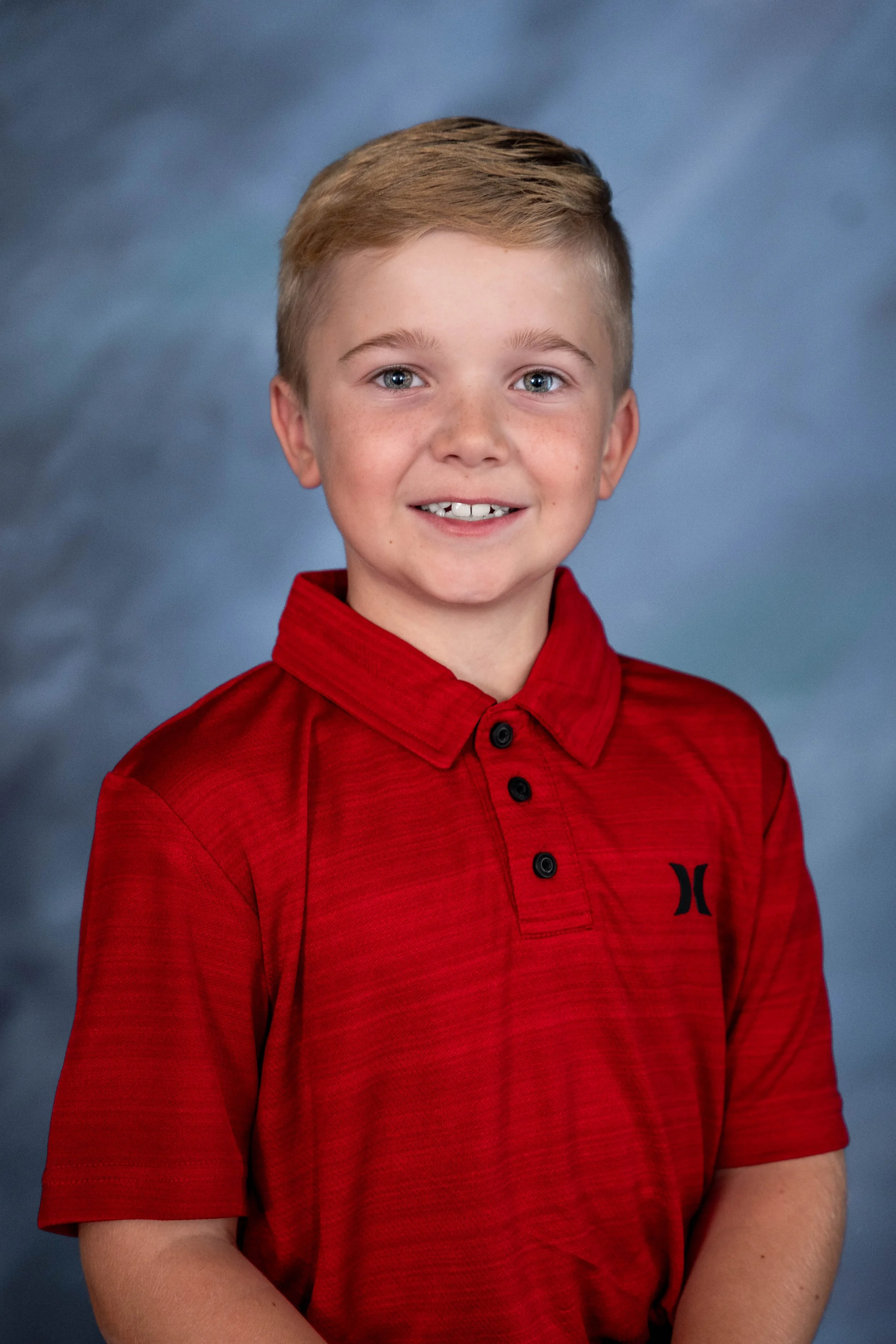 A young boy with blonde hair, blue eyes, and freckles, wearing a red polo shirt, smiling against a blue cloudy backdrop.