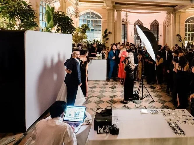 A wedding reception with guests socializing, a photographer setting up photos, and a person working on a laptop at a table, in a decorated hall with large windows and potted plants.