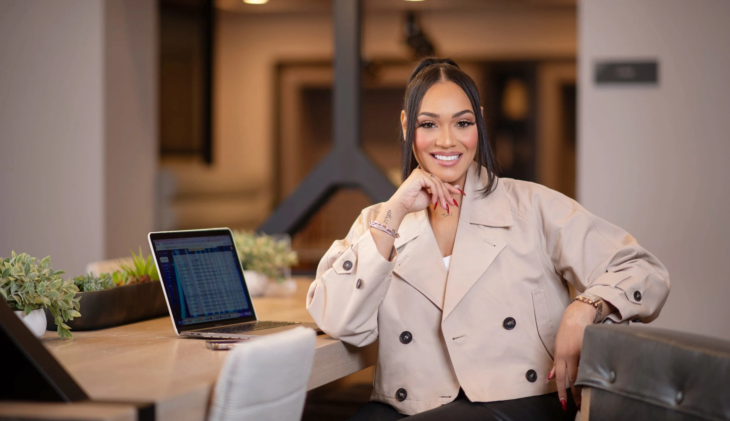 A woman with dark hair tied back, wearing a beige trench coat, sits at a desk in an indoor setting, smiling at the camera. On the desk, there is a laptop displaying a spreadsheet or similar document, a smartphone, and some plants. The background features warm indoor lighting and wooden decor.