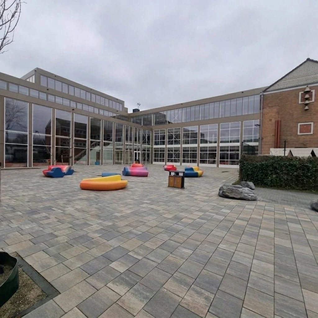 Schoolgebouw met grote ramen en een betegeld schoolplein met kleurrijke zitbanken.