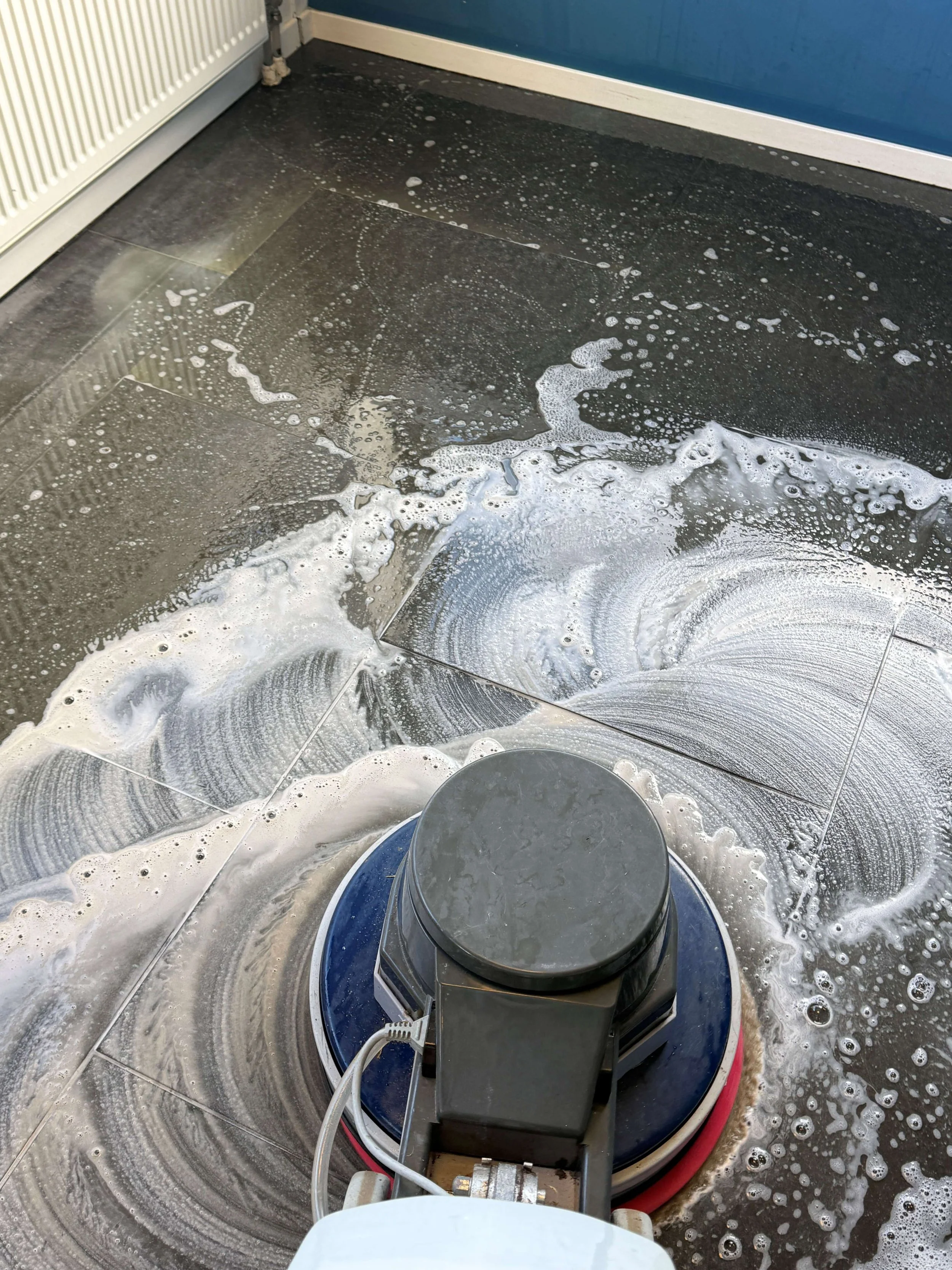 Roterende schrobmachine reinigt een donkere tegelvloer met reinigingsmiddel.