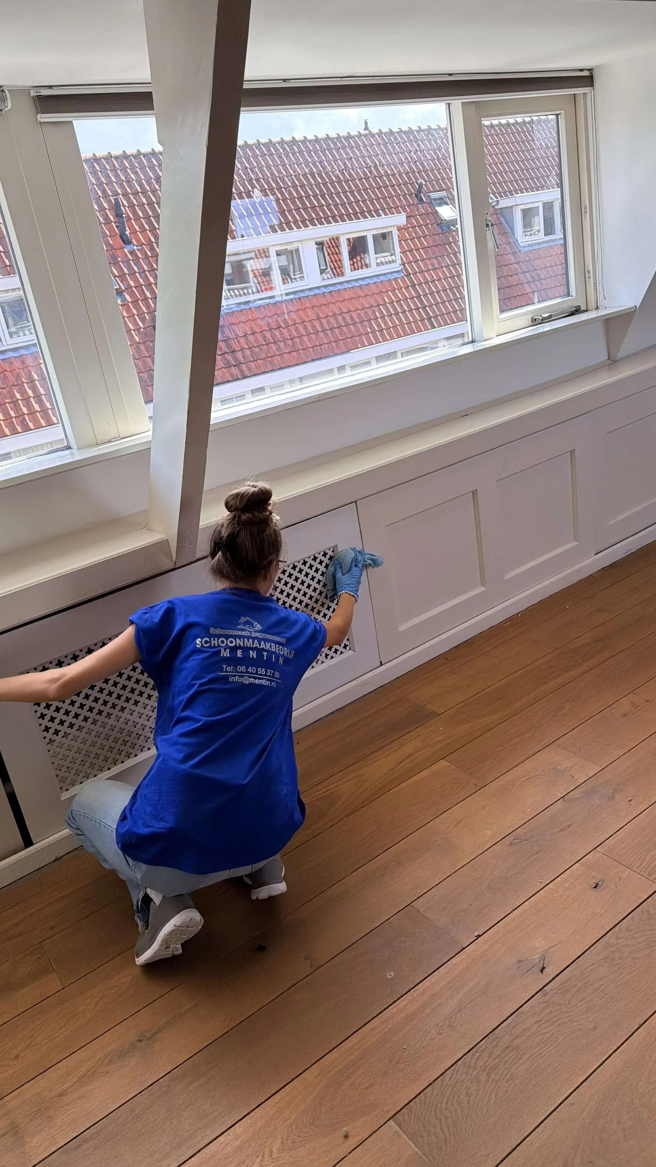 choonmaker in een blauw Mentin-shirt reinigt de radiatorombouw onder een raam.