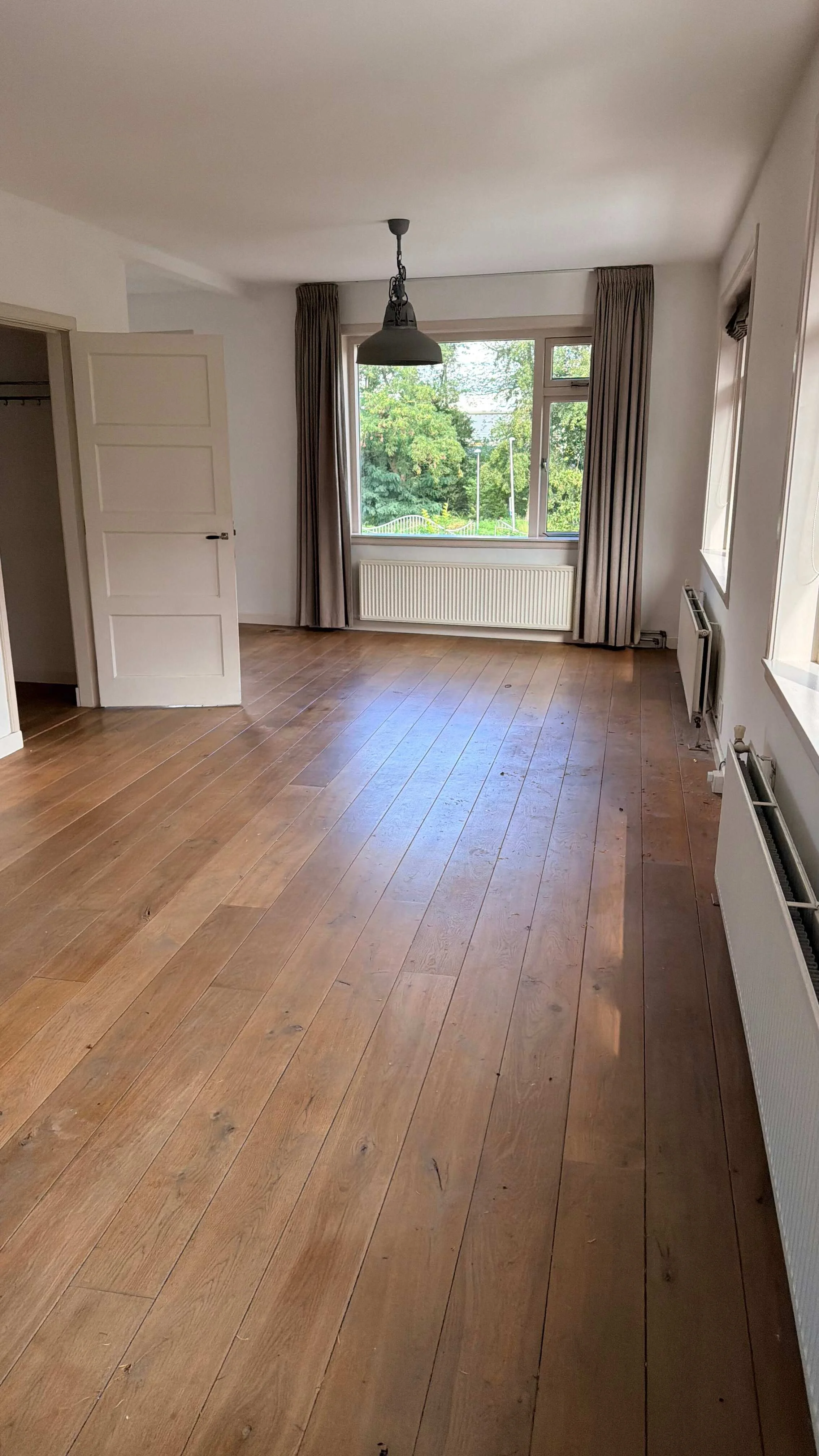 Lege woonkamer met houten vloer, ramen en radiatoren die klaar is voor schoonmaak vóór de verhuizing.