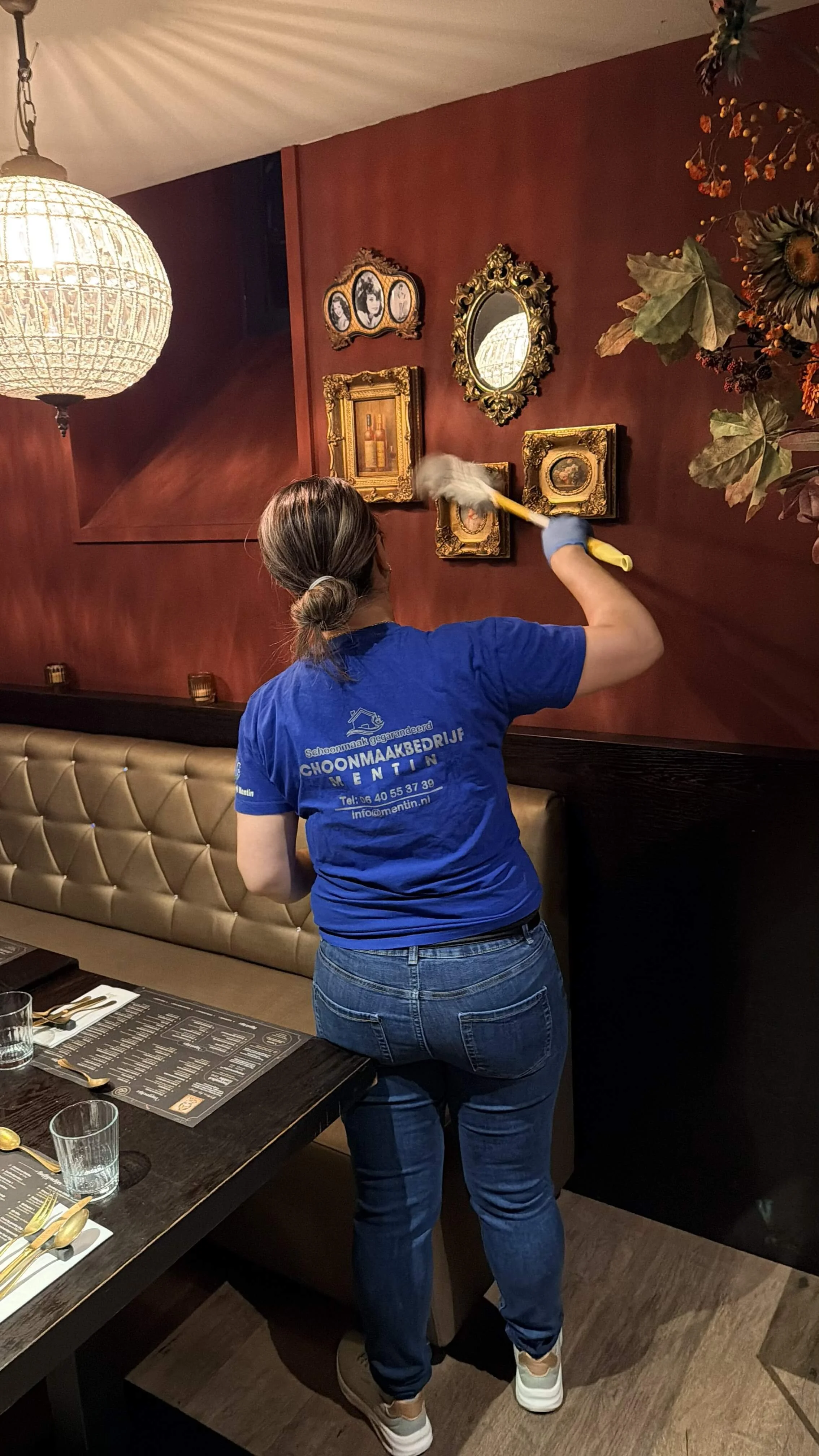 Schoonmaker neemt stof af van wanddecoratie boven een zitbank in een restaurant.