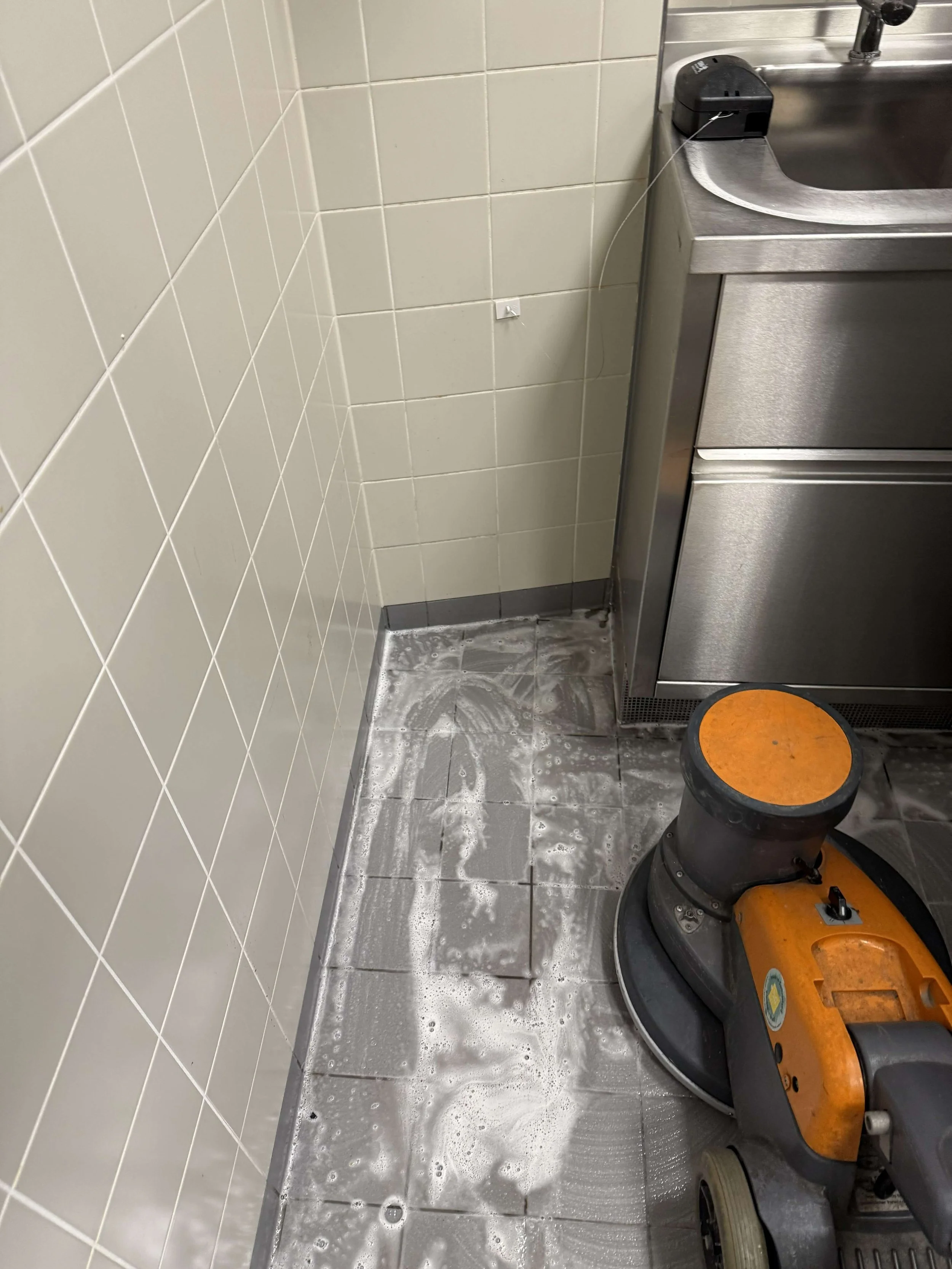 Machinale reiniging van een tegelvloer naast een roestvrijstalen sanitaire voorziening.