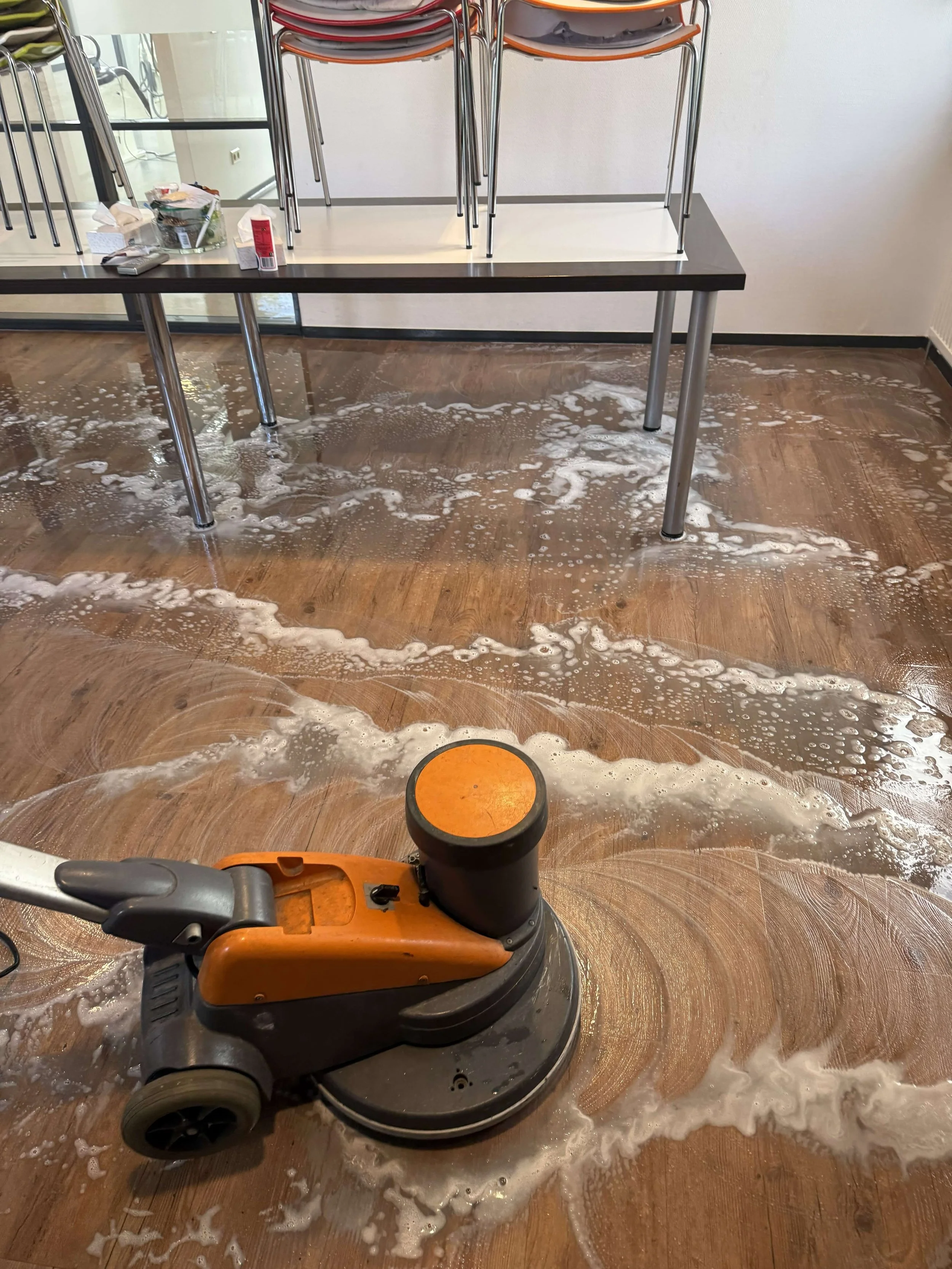Reinigingsmachine schrobt een houten vloer onder een tafel en stoelen.
