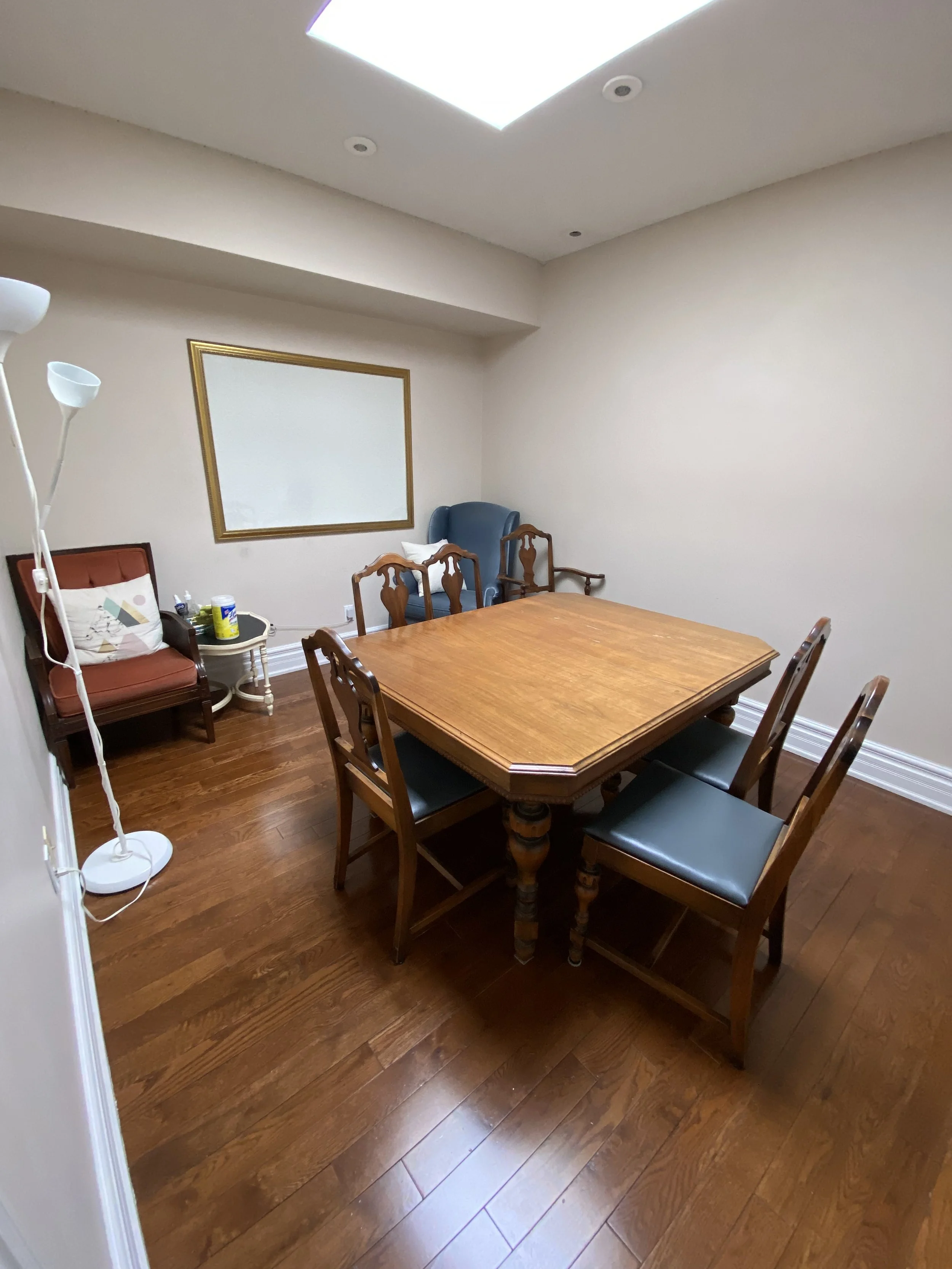 A small conference room with a wooden table and four chairs on a hardwood floor. There is a whiteboard on the wall, a standing lamp, and two additional chairs in the corner.