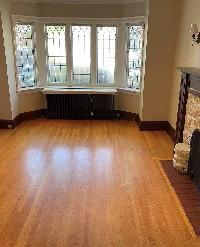 Empty room with hardwood floor, large bay windows with decorative panes, a radiator beneath the windows, and a stone fireplace with a wooden mantel.