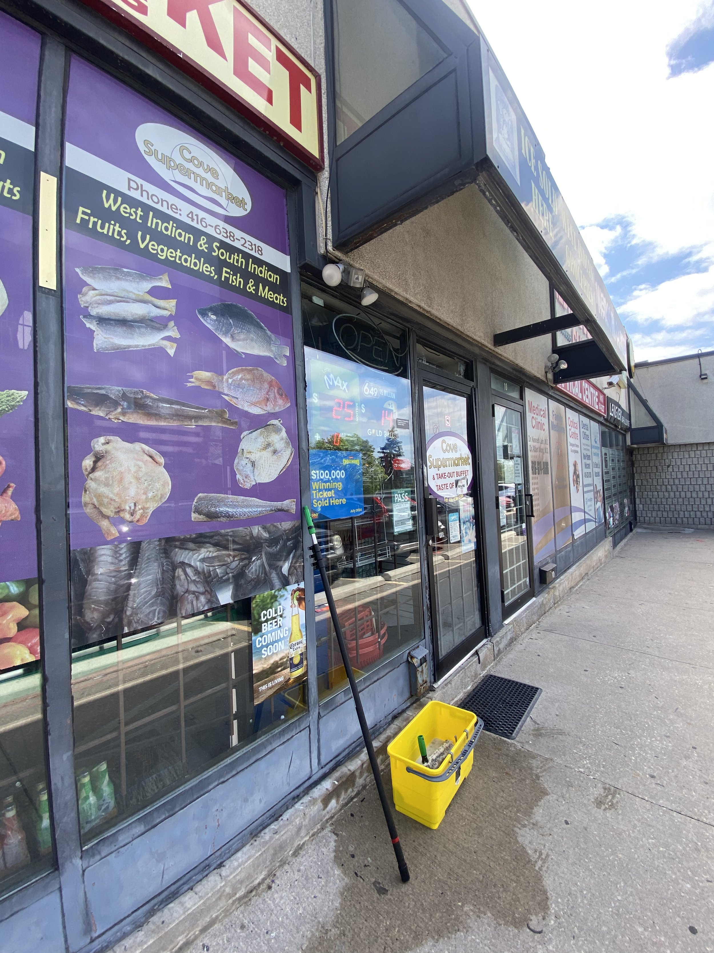 Exterior view of Cowe Supermarket storefront with large purple signs displaying images of fish, fruits, vegetables, and meats. Yellow bucket with cleaning supplies and a broom leaning against the window.