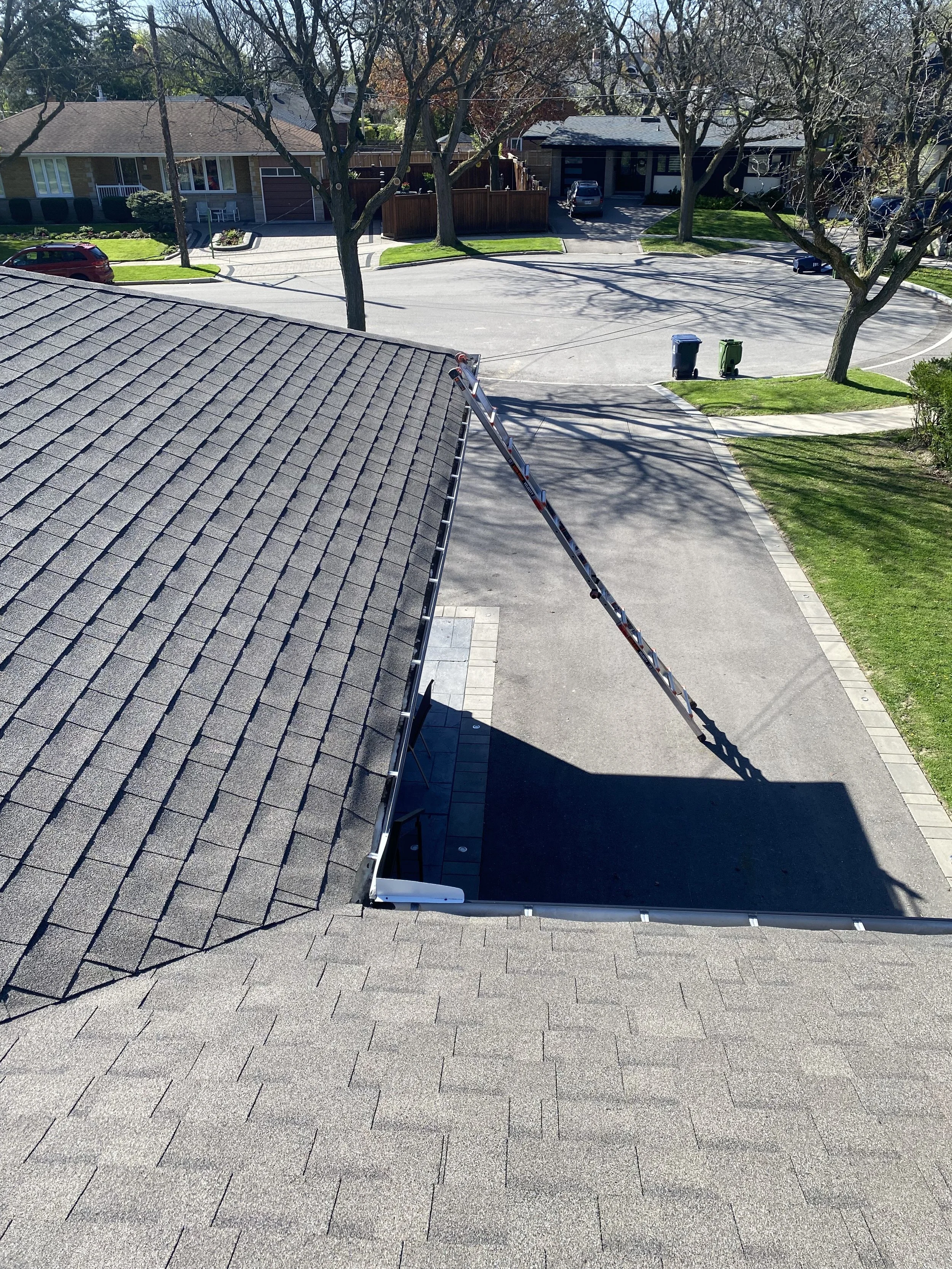 View from a rooftop showing a roof ladder leaning on the shingles, a backyard with trees, a parking lot, and residential houses with driveways.