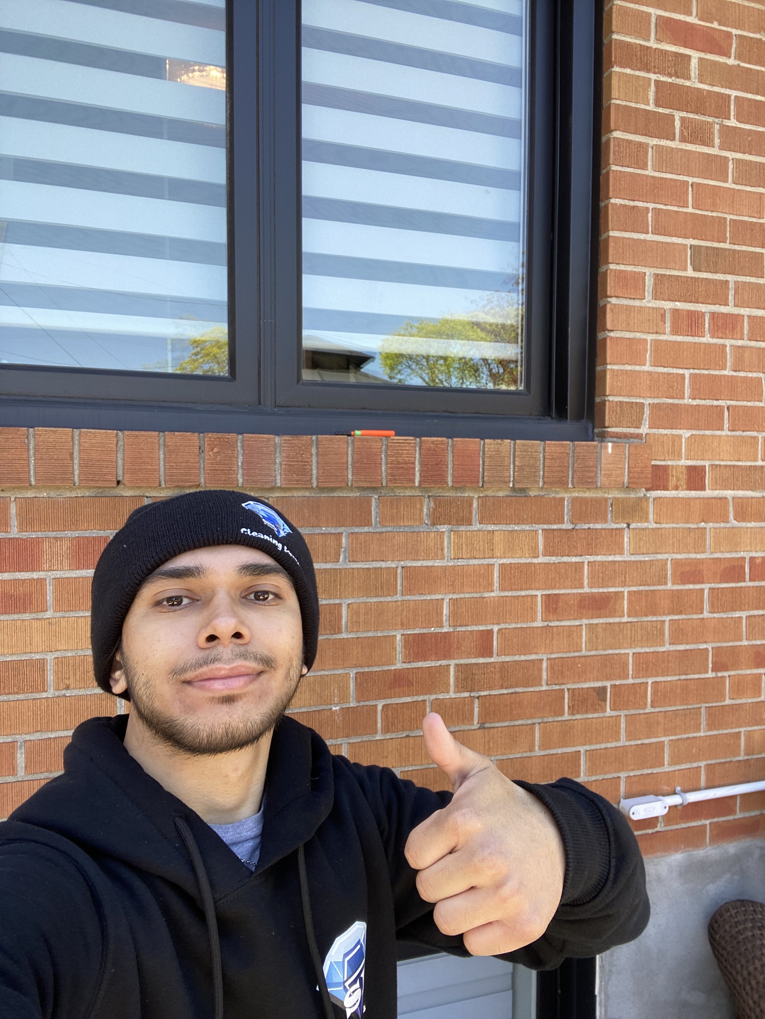 A young man taking a selfie outside a brick building, wearing a black hoodie and a black beanie, giving a thumbs-up gesture.
