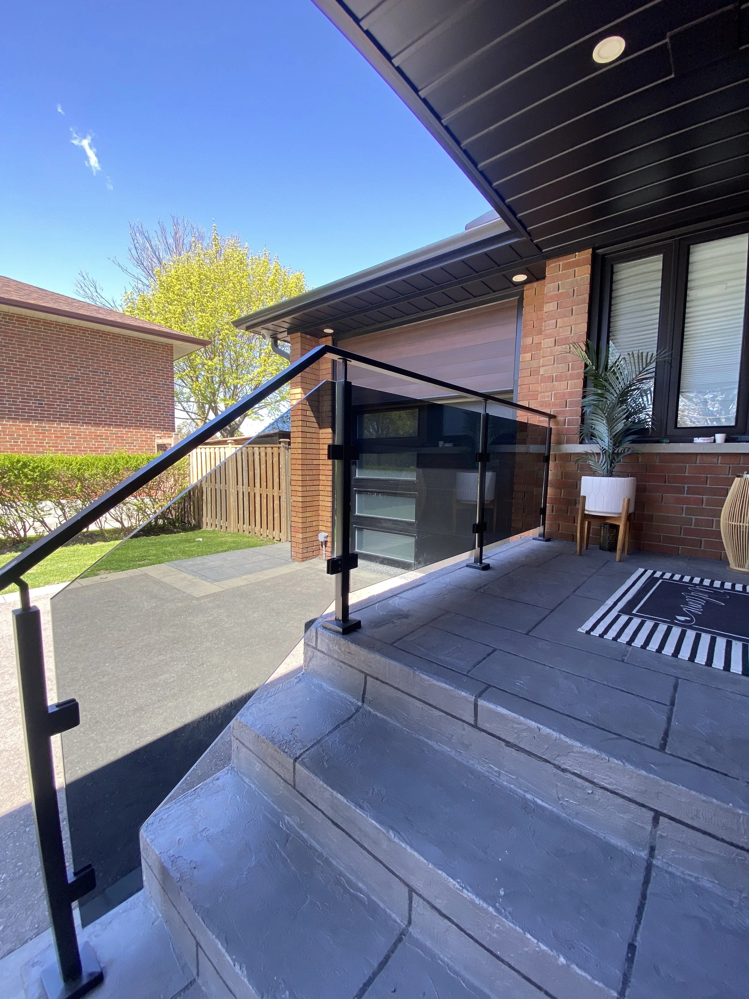 A front porch with stone steps, black metal and glass railing, a welcome mat, potted plant, and brick exterior of a house under a blue sky.