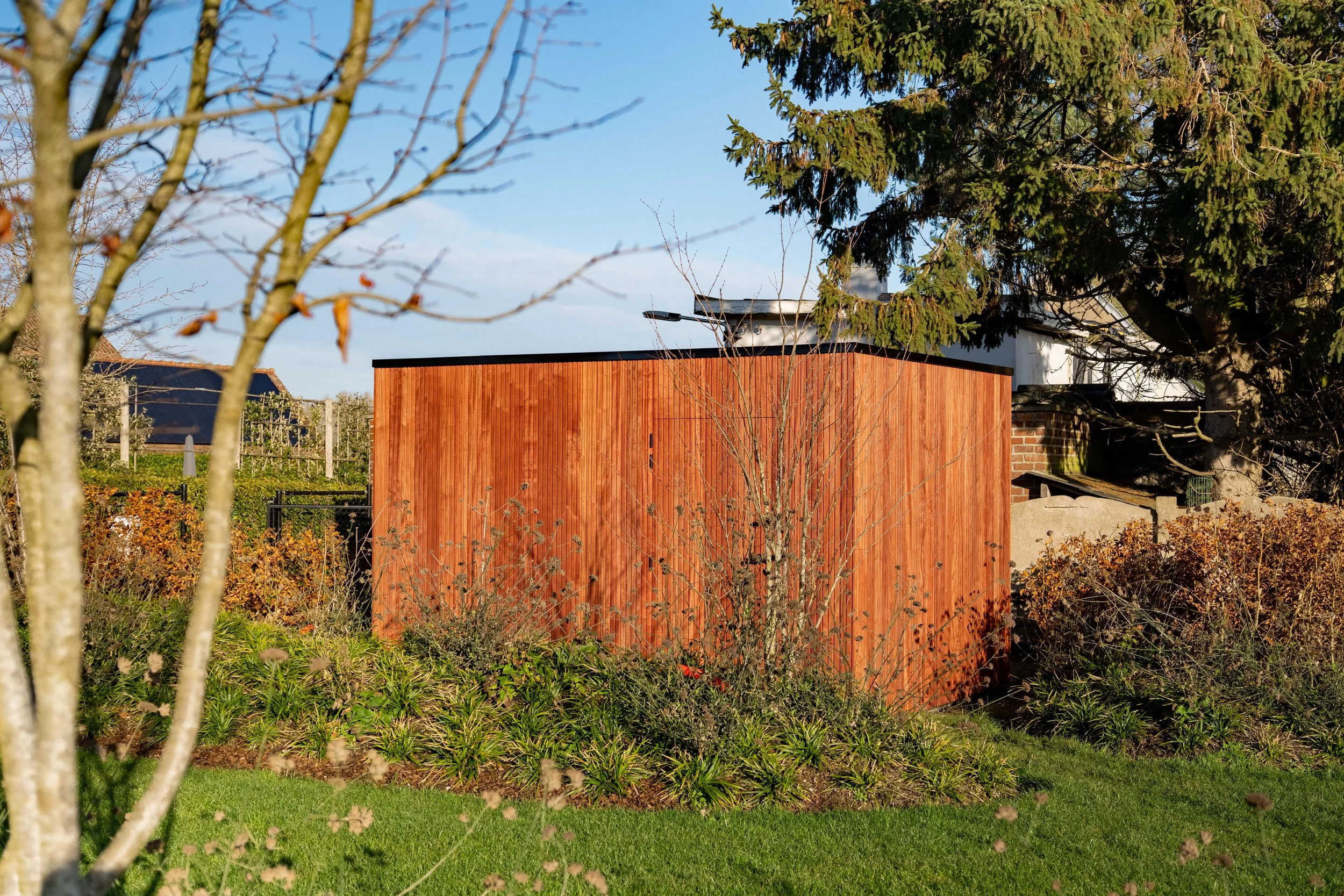 Een tuin met een houten tuinhuis, bomen en struiken, zonlicht en een blauwe lucht