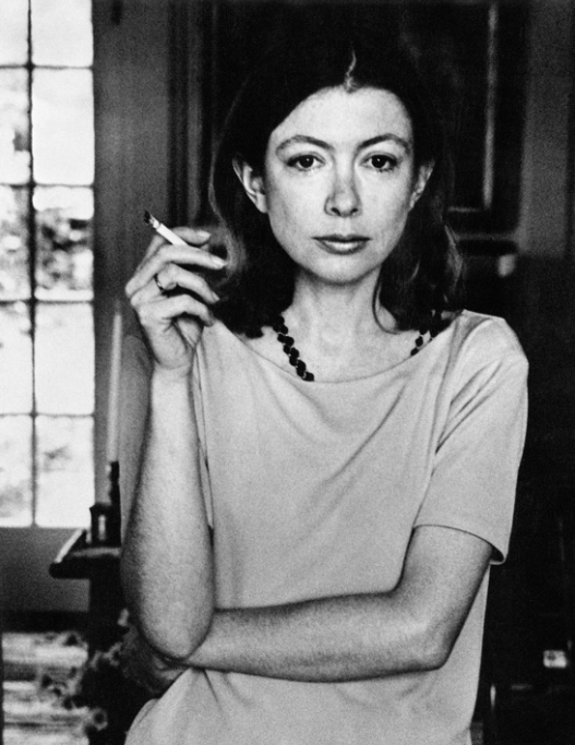 A black and white portrait photograph of Joan Didion. She is holding a lit cigarette with one arm crossed