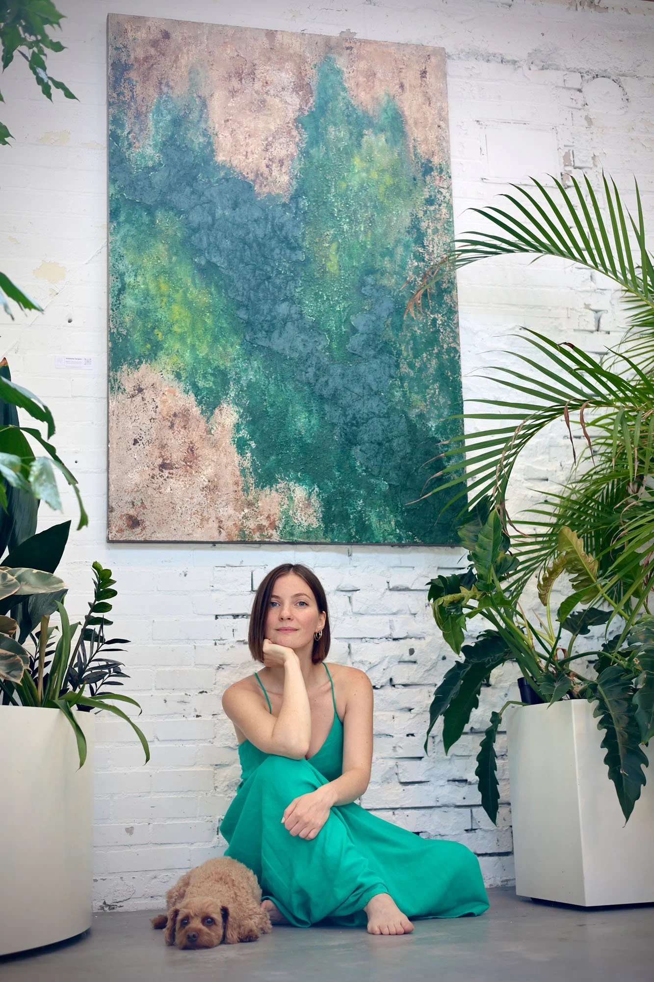 A woman with short brown hair sitting on the floor in front of a white brick wall, surrounded by large potted plants. She is wearing a turquoise dress and has her chin resting on her hand, with a gentle smile. A small tan-colored dog is lying on the floor in front of her. There is a colorful abstract painting hanging on the wall behind her.