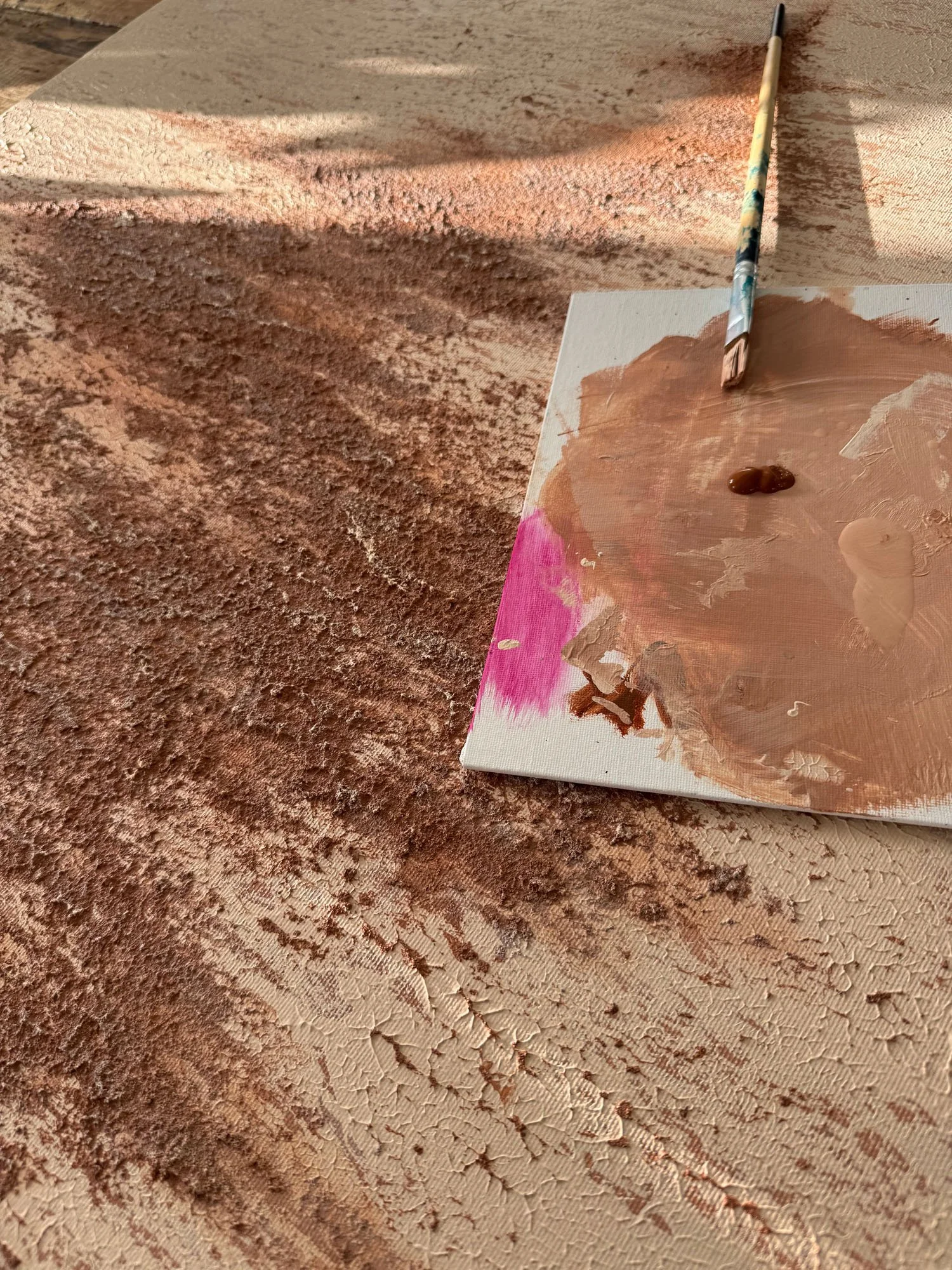 Paint palette with beige, pink, and brown paints, and a paintbrush resting on it, resting on a textured surface with dry, cracked paint.