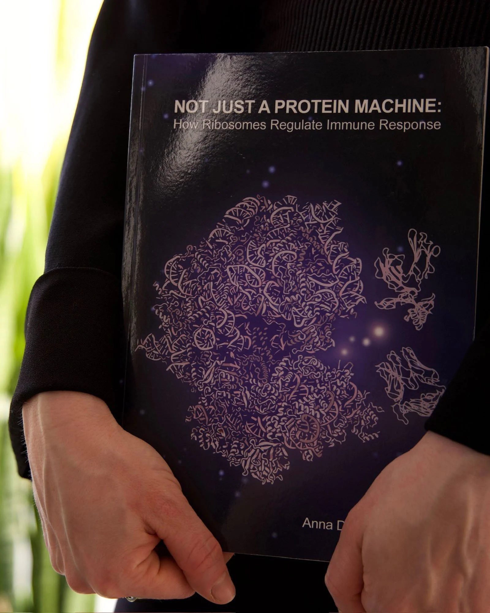 Close-up of a person holding a book titled 'NOT JUST A PROTEIN MACHINE: How Ribosomes Regulate Immune Response' by Anna D. The person's hand is gripping the lower part of the book, and they are wearing a black sleeve. The background appears to be a green environment, possibly outdoors.