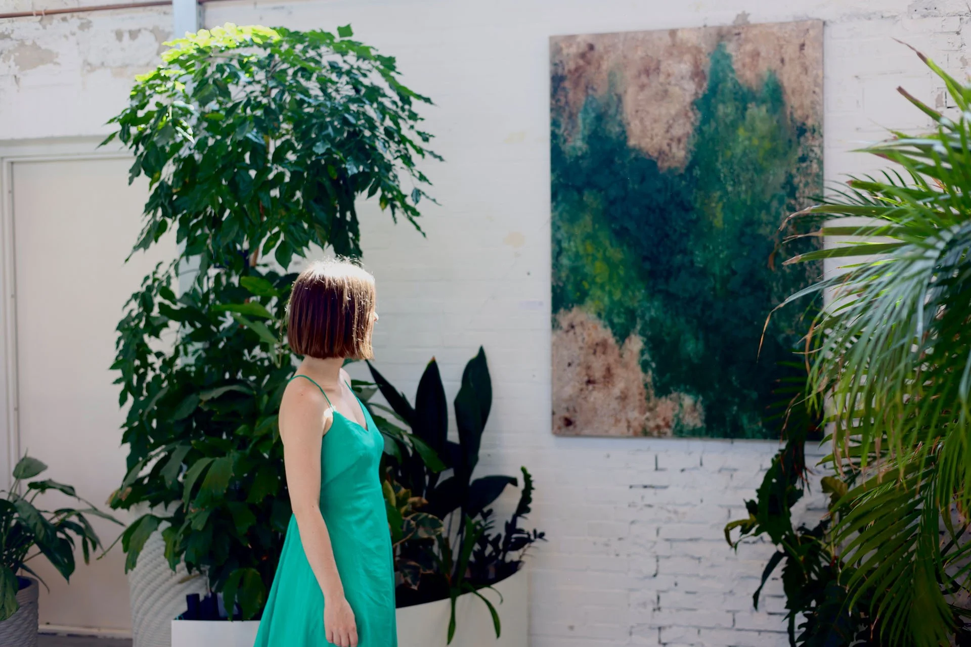 A woman in a green dress standing among large potted plants and greenery, looking at an abstract painting on a white brick wall.