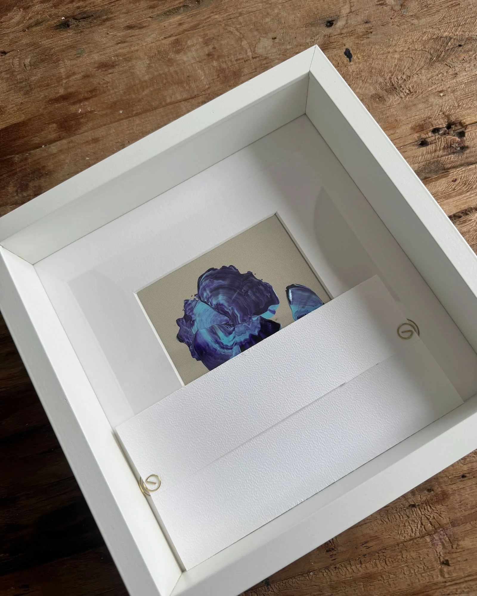 A white picture frame leaning against a wooden surface, with a painting or print visible inside the frame showing abstract blue and purple shapes.