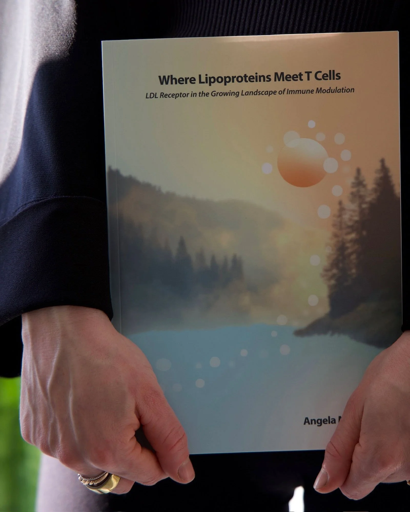 Close-up of a person holding a book titled "Where Lipoproteins Meet T Cells: LDL Receptor in the Growing Landscape of Immune Modulation" with a scenic landscape background on the cover.
