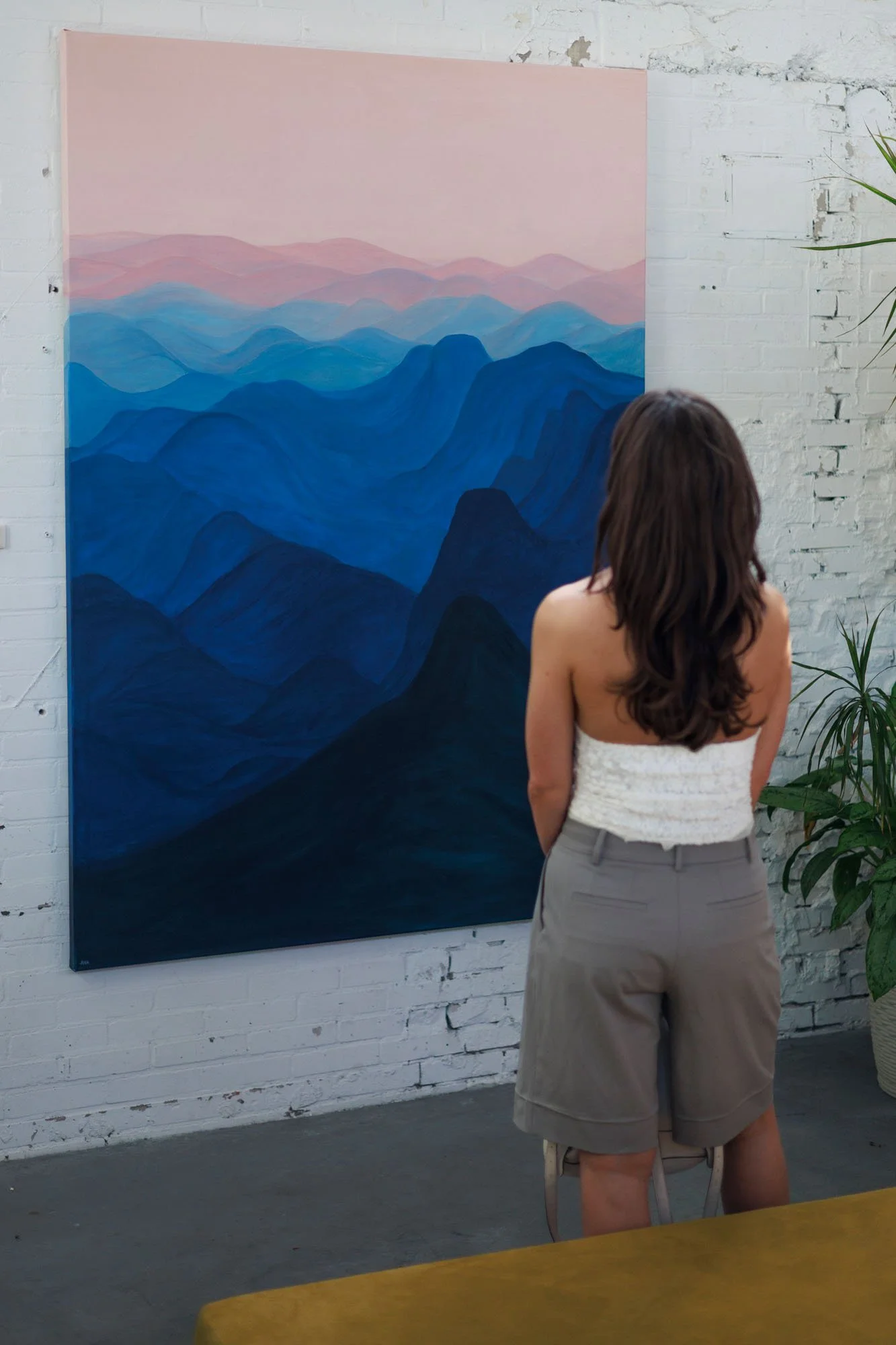 A woman with long brown hair, wearing a strapless white top and beige shorts, is standing and looking at a large painting of layered mountains in pastel pinks and blues on a white brick wall in an art gallery.