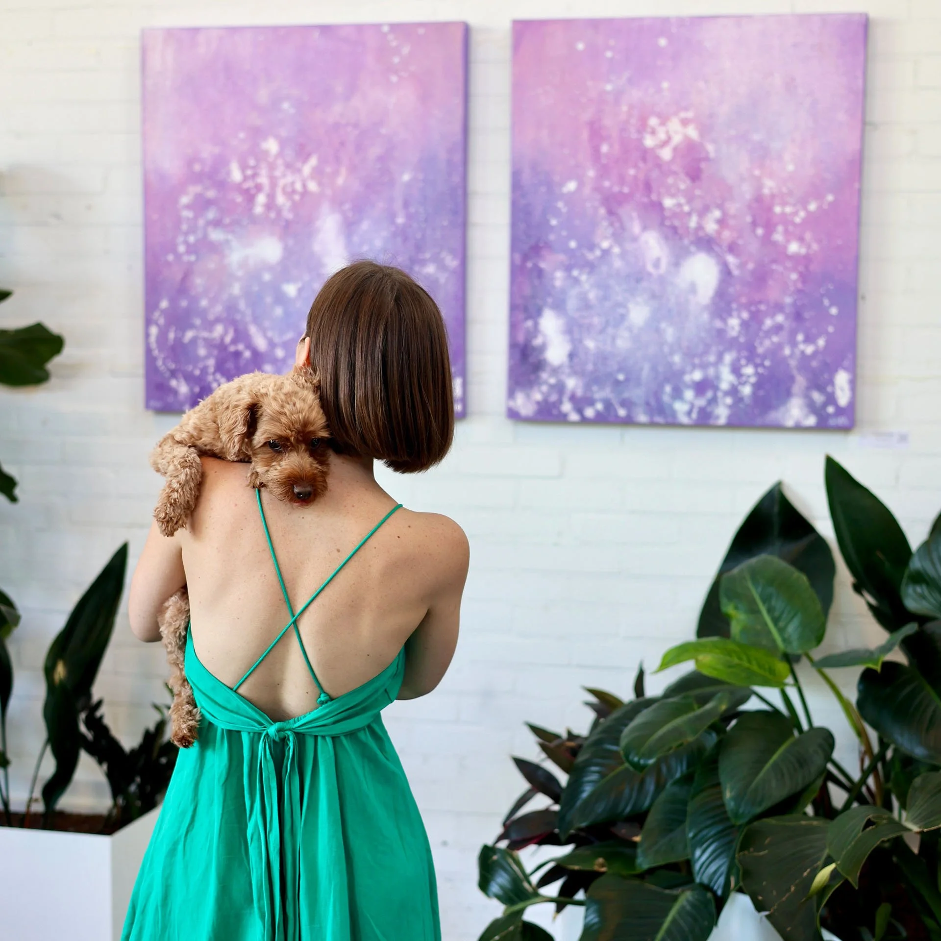 A woman with brown hair holding a small brown poodle puppy. She is wearing a teal dress with spaghetti straps and is facing away from the camera. Behind them are two purple abstract paintings on a white wall, and there are large green houseplants in the foreground.