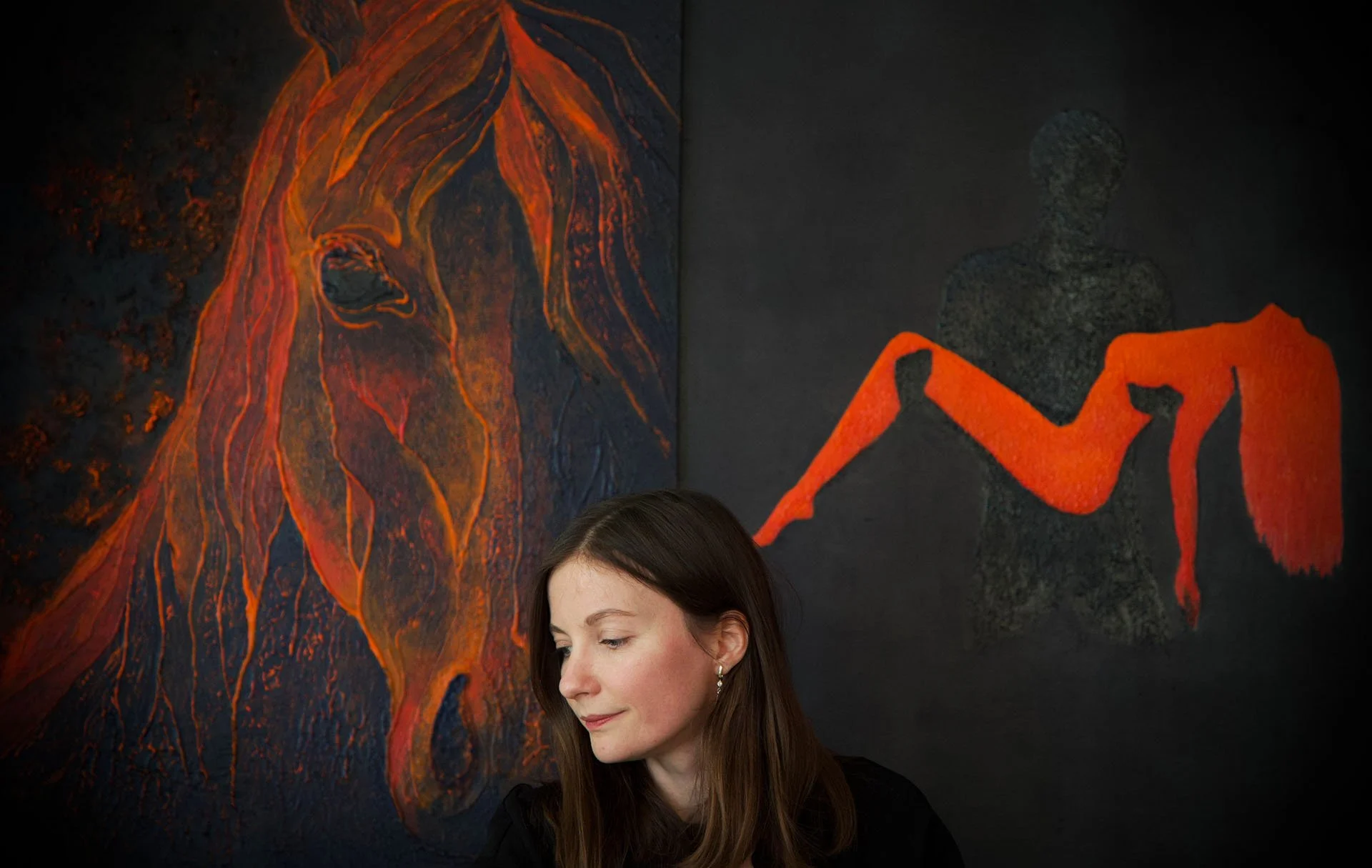A woman with brown hair standing in front of a black wall with artwork of a horse's face and a reclining human figure.