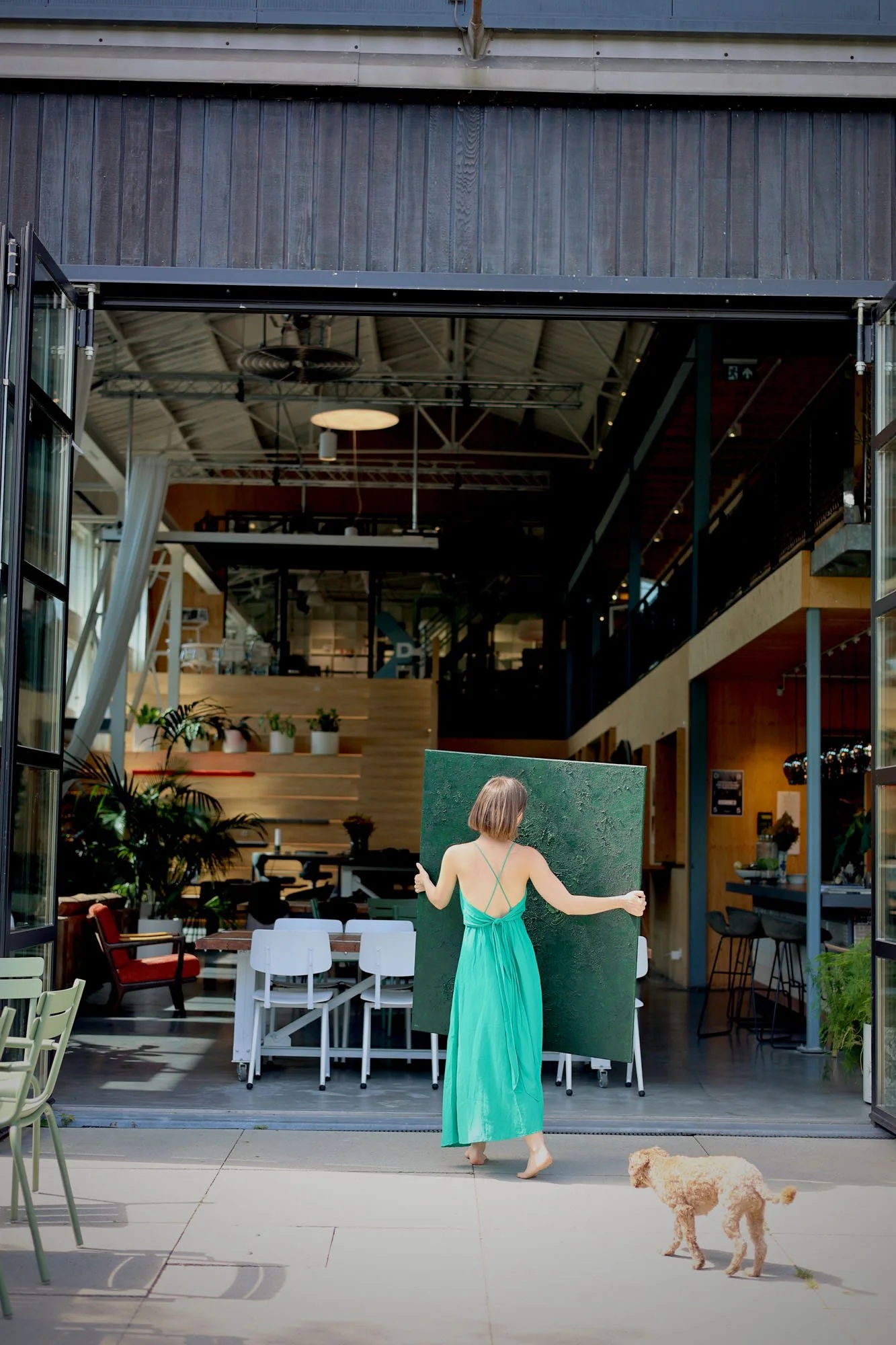 A woman in a green dress is standing barefoot on a patio, facing away while holding a large green board. A small dog is nearby, walking on the patio. The background shows an indoor space with modern furniture, plants, and a high ceiling with exposed beams.