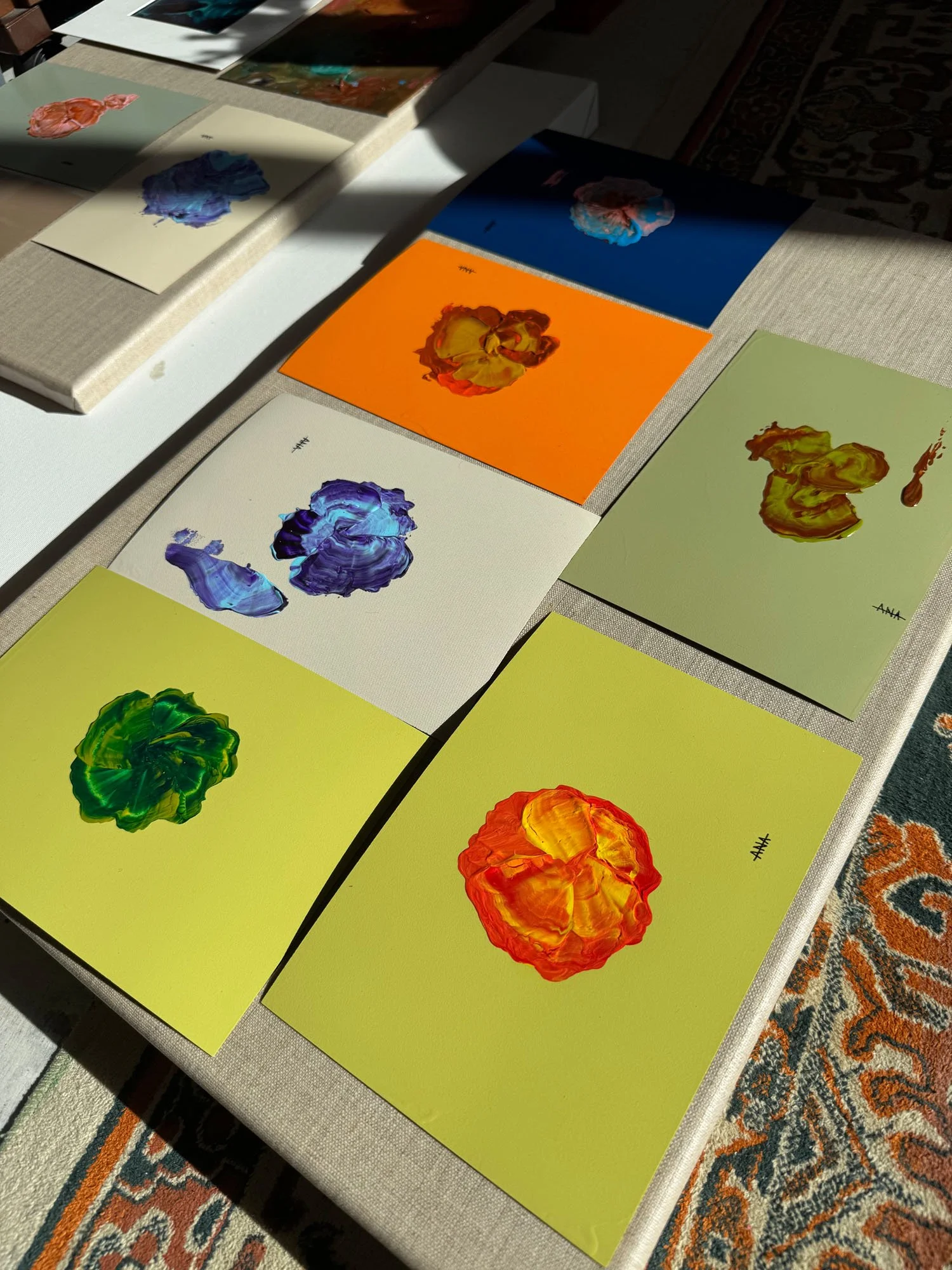 Colorful abstract paintings arranged on a table, with sunlight casting shadow across some of them.