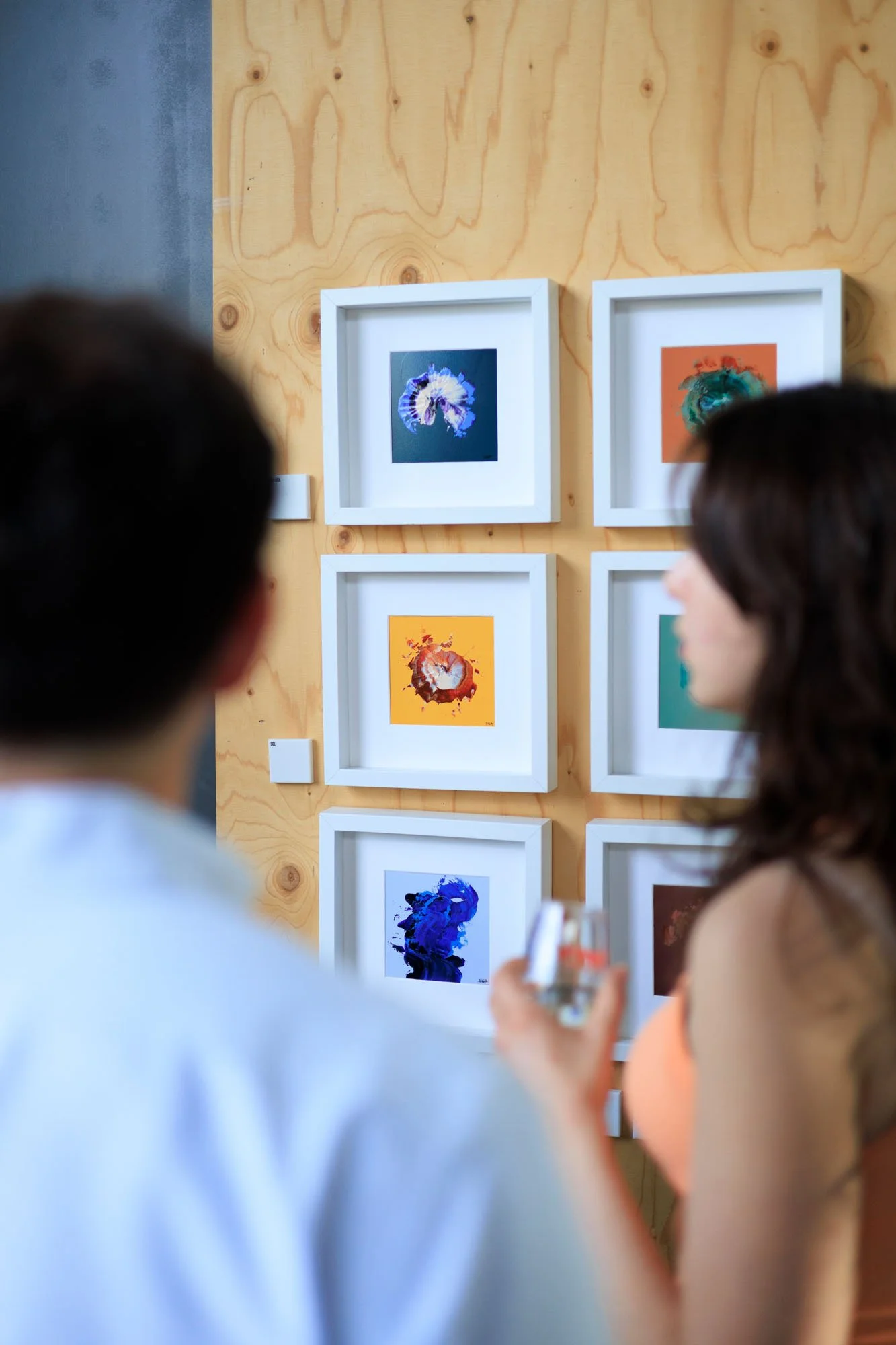 People viewing artwork in a gallery with framed colorful abstract paintings on a light wooden wall.