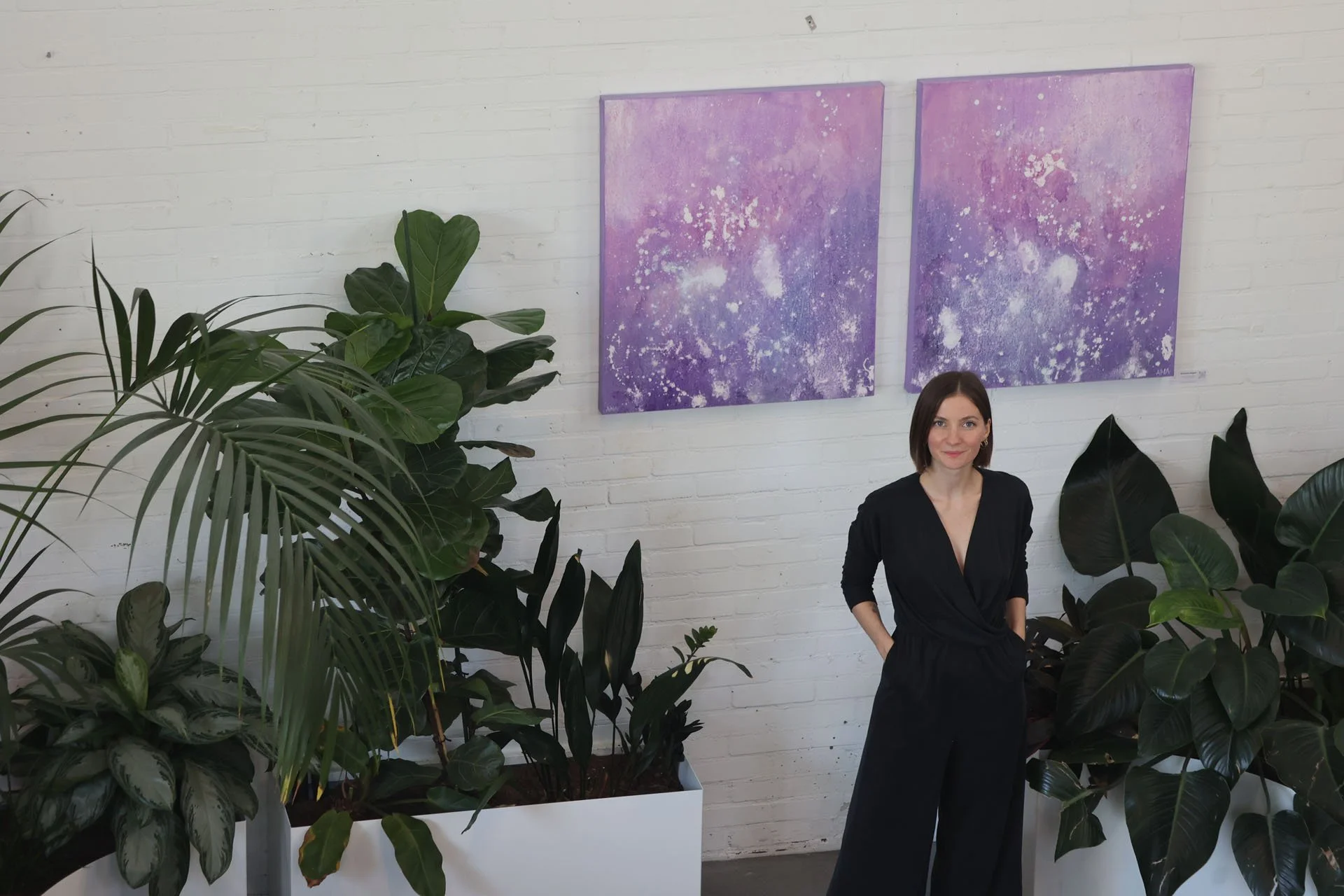 A woman in a black outfit stands surrounded by large green indoor plants in front of a white brick wall with two purple and white abstract paintings.