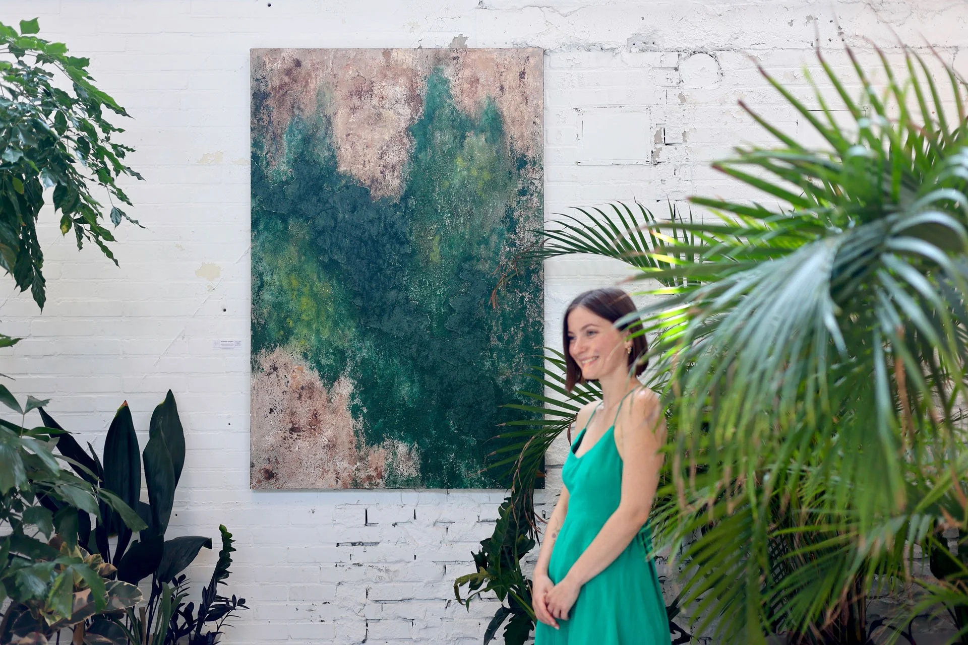 A woman with short brown hair in a teal dress standing among green plants in a room with white brick walls and a large abstract painting.