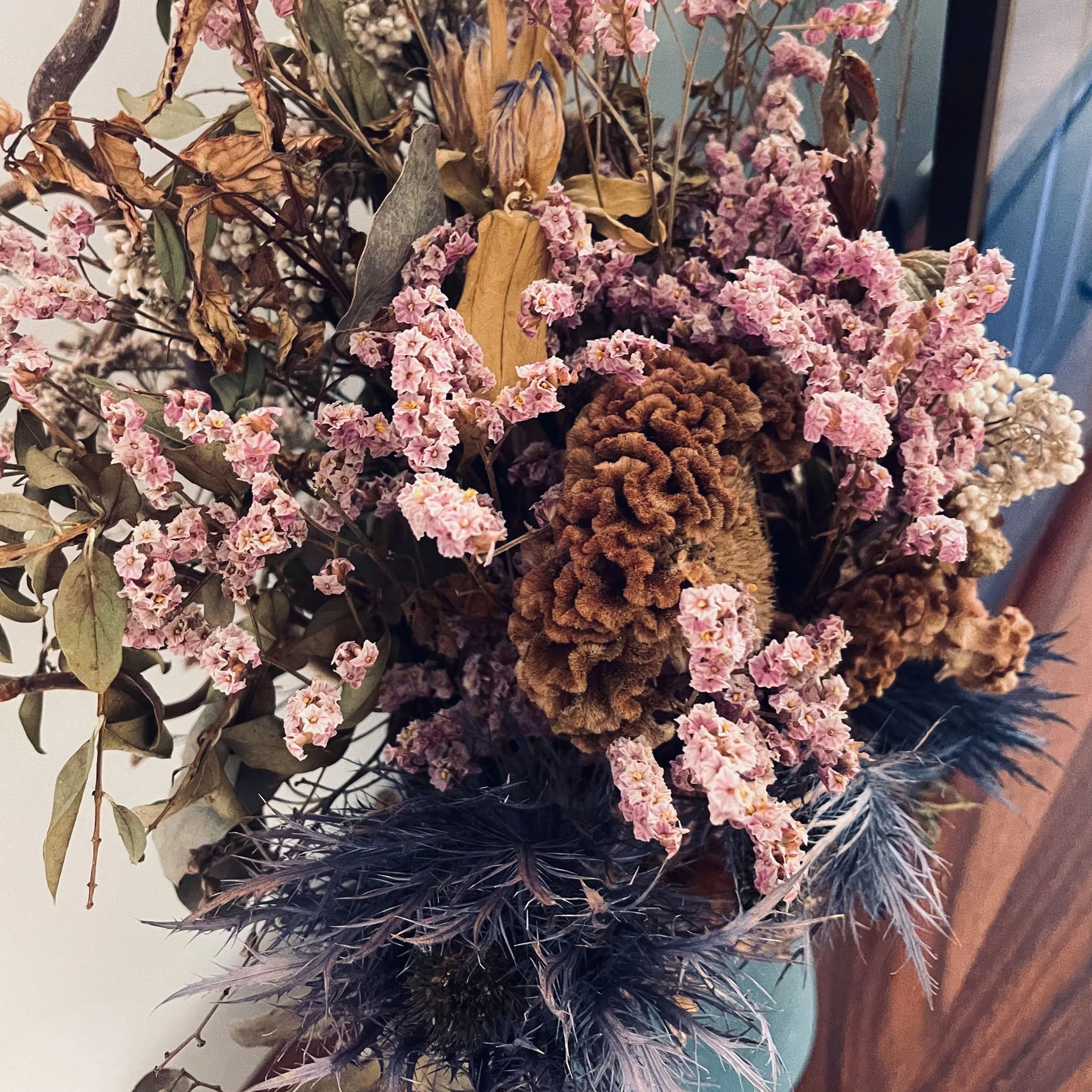A close-up view of a dried floral arrangement featuring pink flowers, brown pine cones, and dark sprigs on a flat surface.