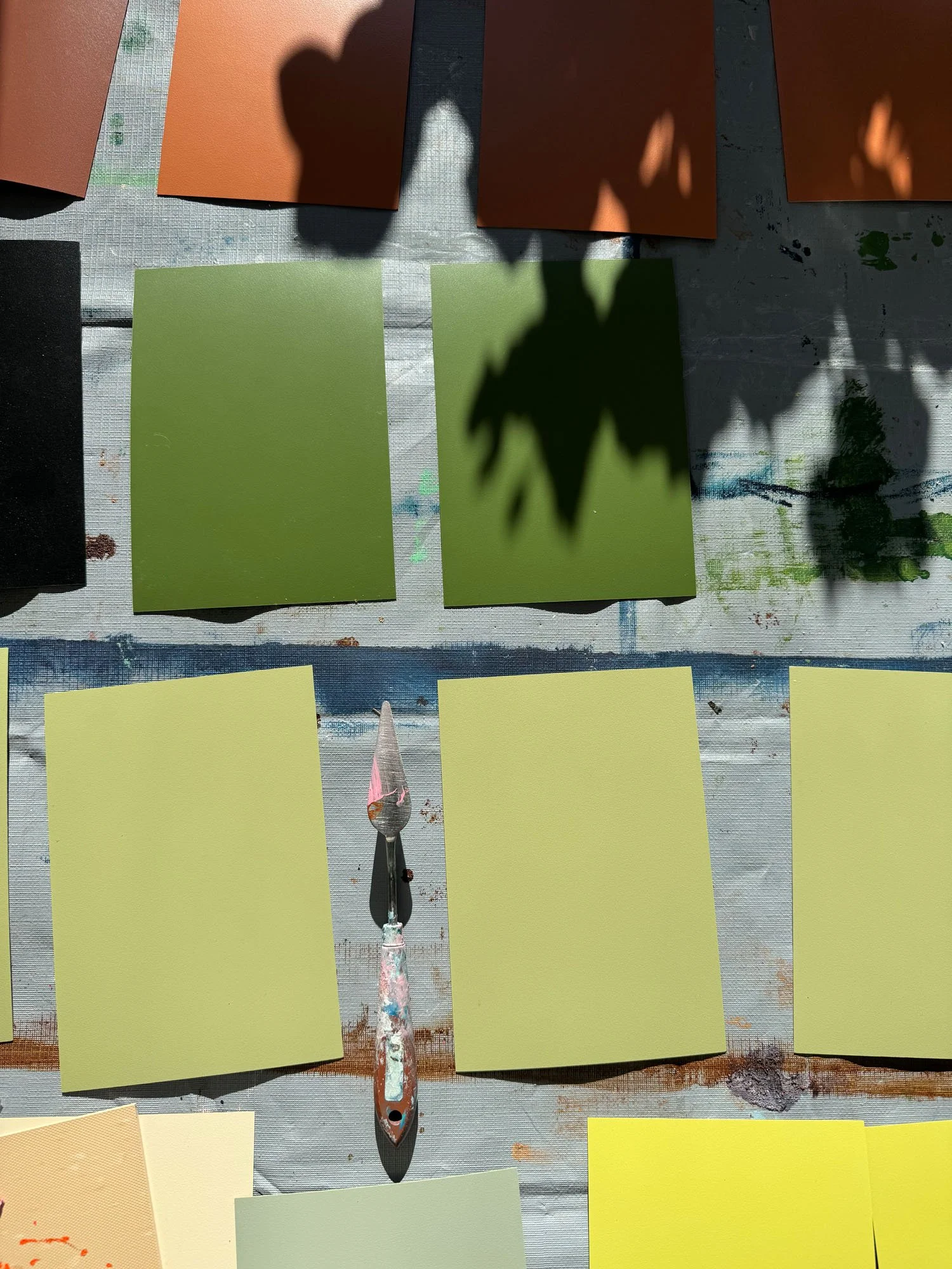 Color swatches in various shades of green, brown, black, and yellow arranged on a surface, with a paintbrush and palette knife in the center, likely part of an art or painting project.
