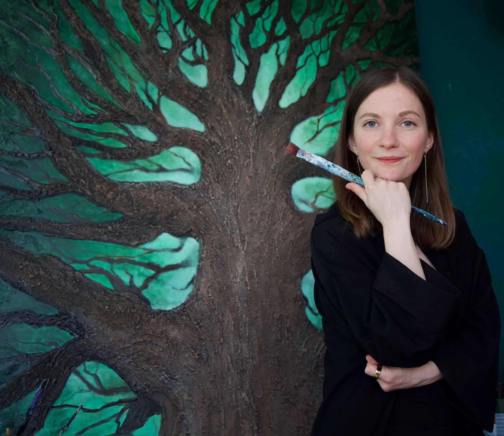 A woman with brown hair, blue eyes, earrings, and a black top holds a paintbrush, standing in front of a large mural of a tree painted on a wall with green leaves and brown bark.