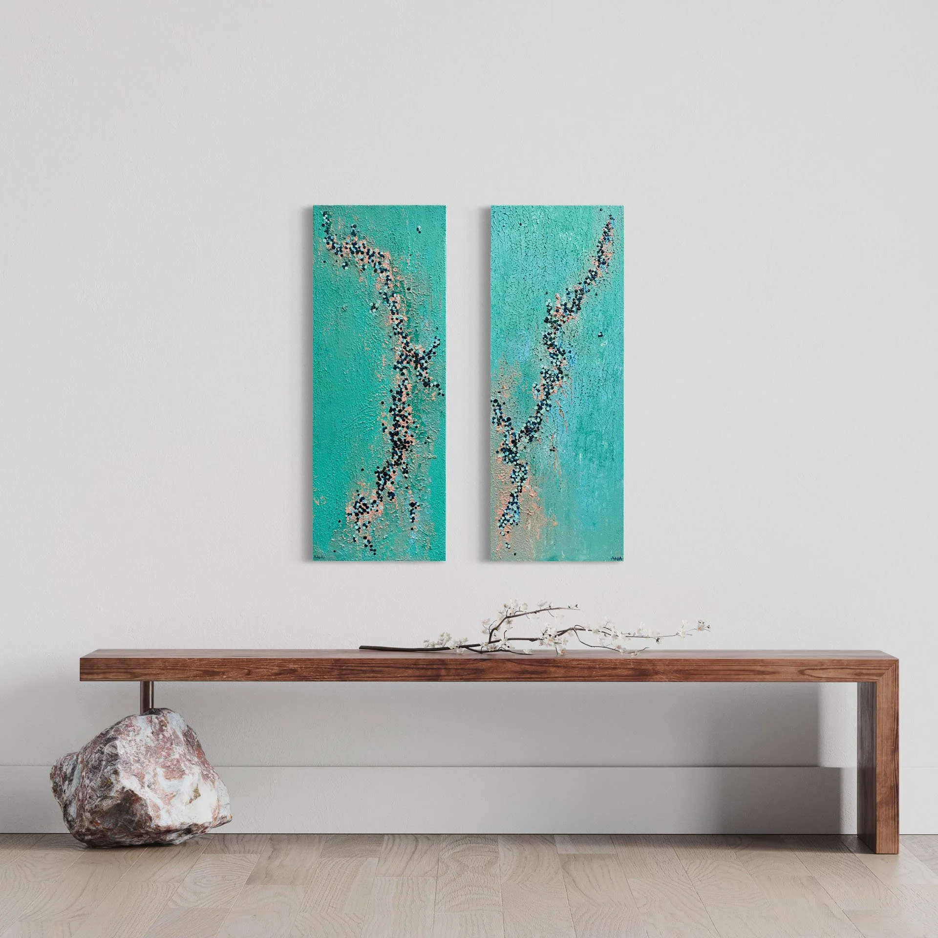 A modern interior with a wooden console table decorated with a branch of white blossoms and a large rock. Above, two abstract paintings on a plain wall feature shades of blue and black dots forming organic patterns.