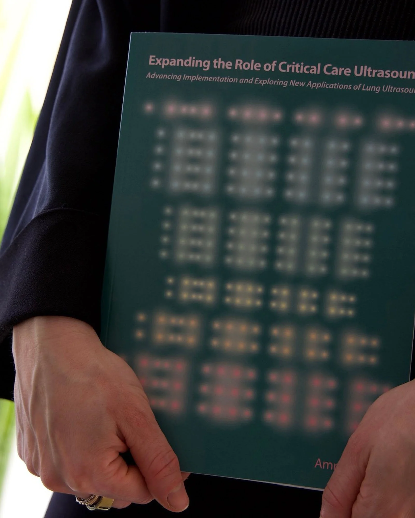 Close-up of a person holding a teal-colored conference or scientific publication titled 'Expanding the Role of Critical Care Ultrasound,' with a subtitle about lung ultrasound applications. The person is wearing a black sleeve and has a gold ring on their finger.
