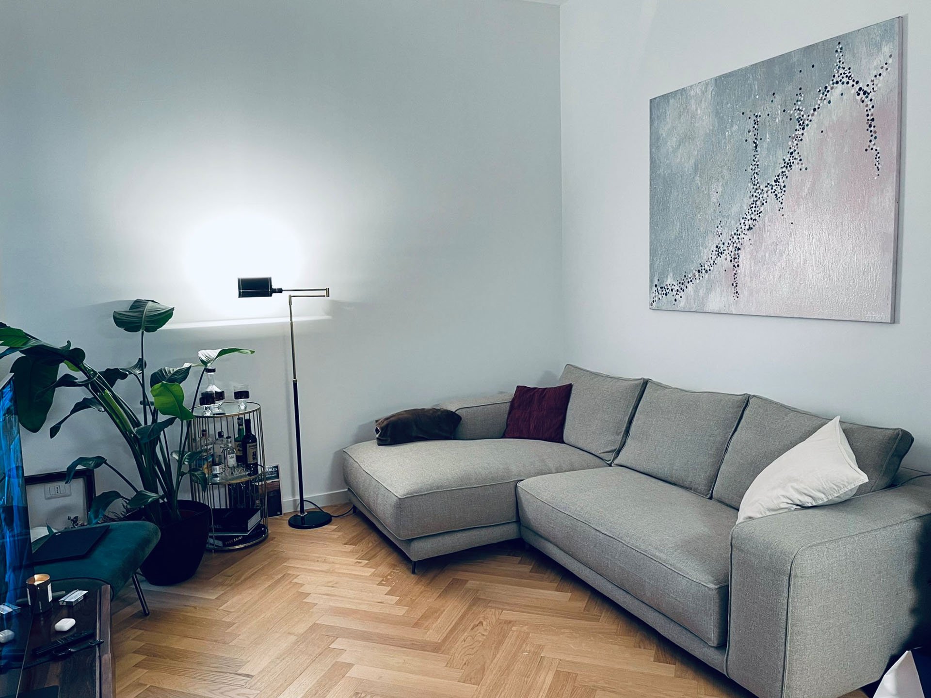 Living room with a light gray sectional sofa, a white pillow, and a purple pillow. Wall art depicting an abstract horse made of small black dots. Floor lamp and a small bar cart with bottles on the left side. Houseplants nearby. Hardwood floor.
