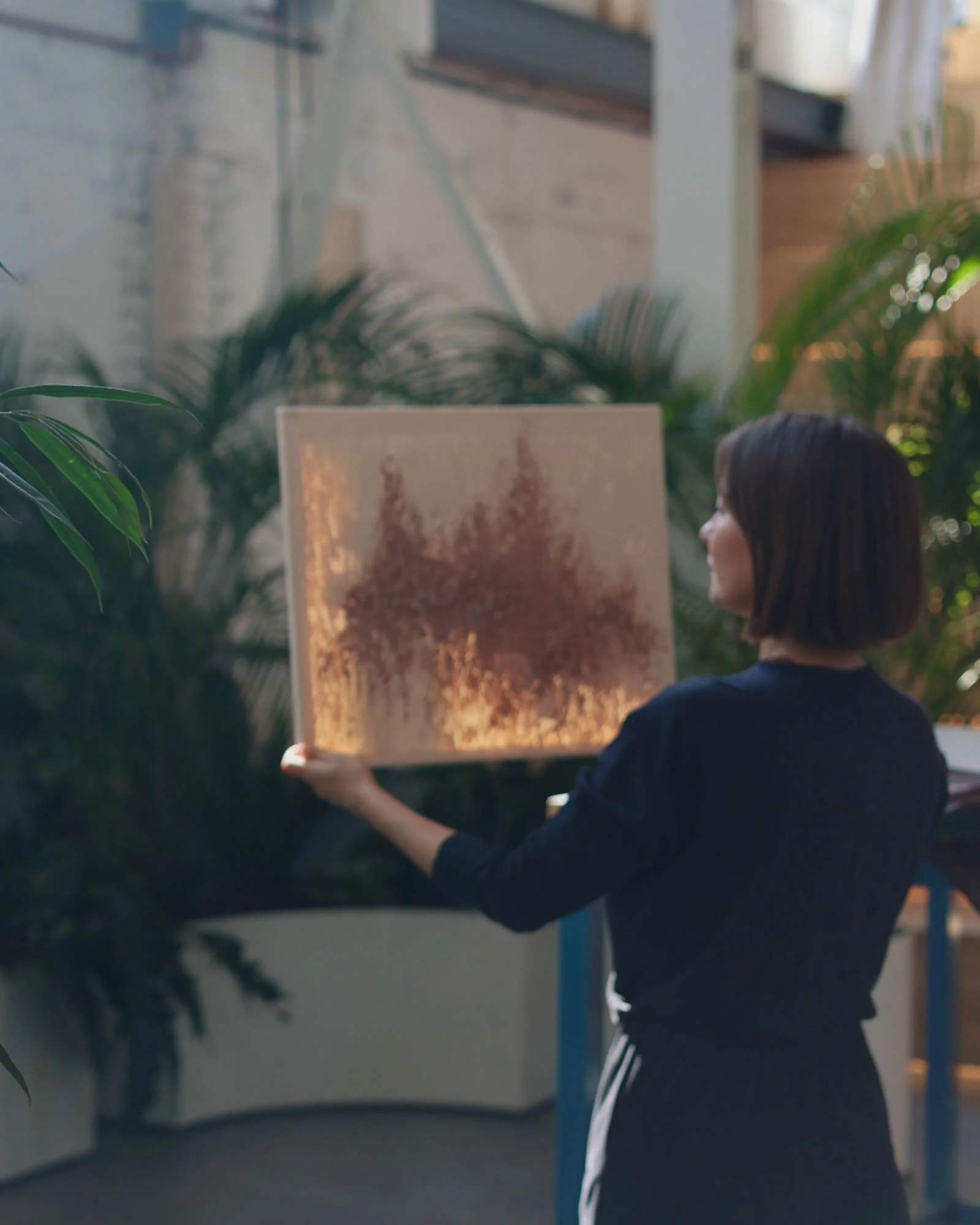 A person with short dark hair wearing a black jacket and white-striped pants, holding a framed picture or artwork in an indoor space with plants and large windows.