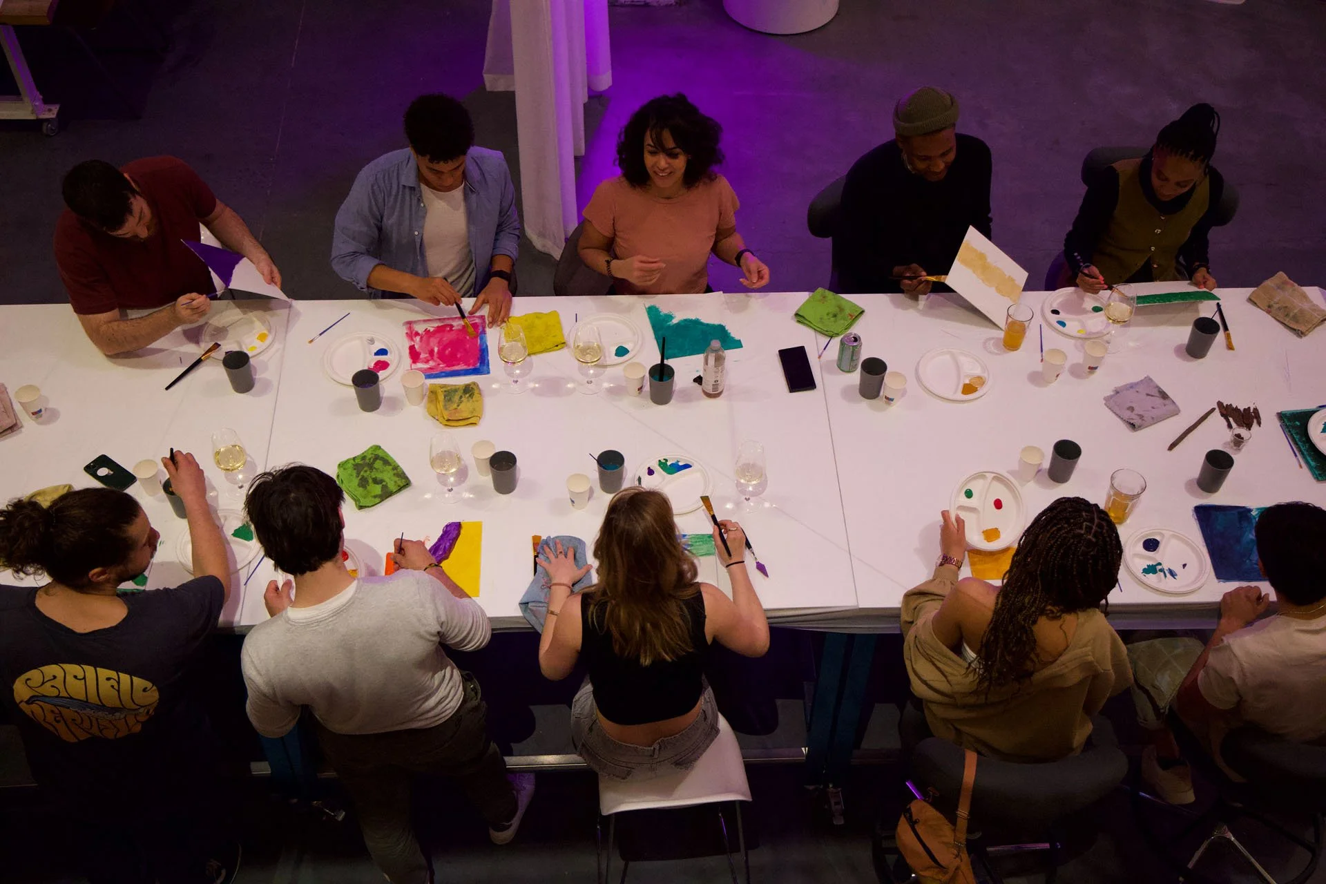 A group of people sitting around a long table painting on canvases, with various paint supplies and drinks on the table. The scene is illuminated with purple lighting.