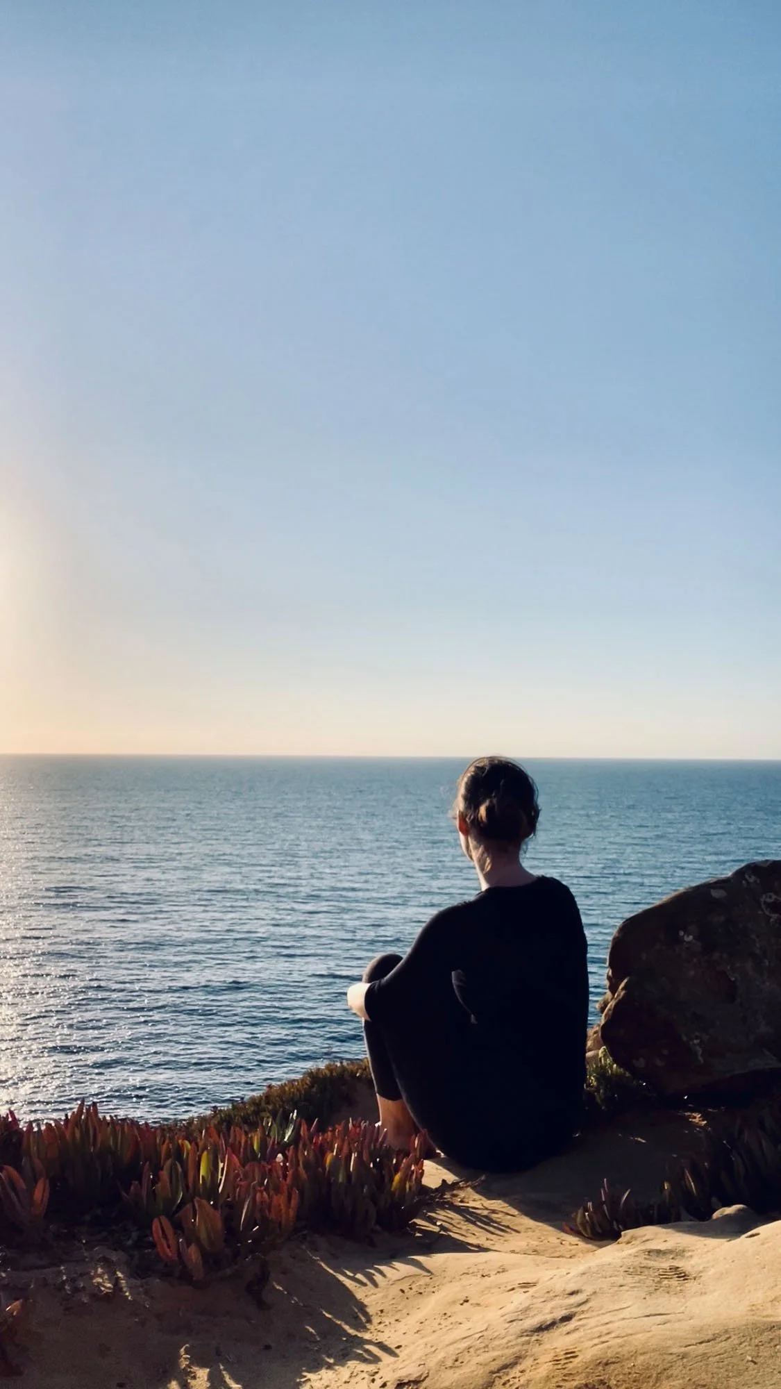 A person sitting on a rocky cliff, looking out at the ocean during sunset.